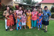 Standing on a lawn in front of a building with lush greenery and tropical plants in the background, TaRanda’s multi-generational family in colorful Hawaiian attire smiles for the camera.