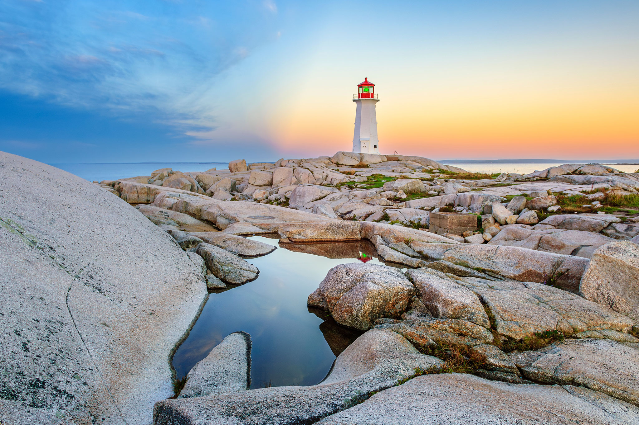 A lighthouse with a white body and red top sits near the ocean’s edge at sunset, with various-sized rocks and small pools of water leading the path toward it.
