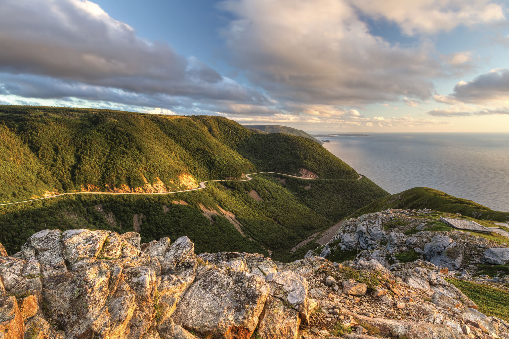 A winding trail cuts through the green cliffs of Cape Breton Highlands National Park, with an incredible view of the rocky shoreline shadowed by puffy clouds and endless miles of ocean.