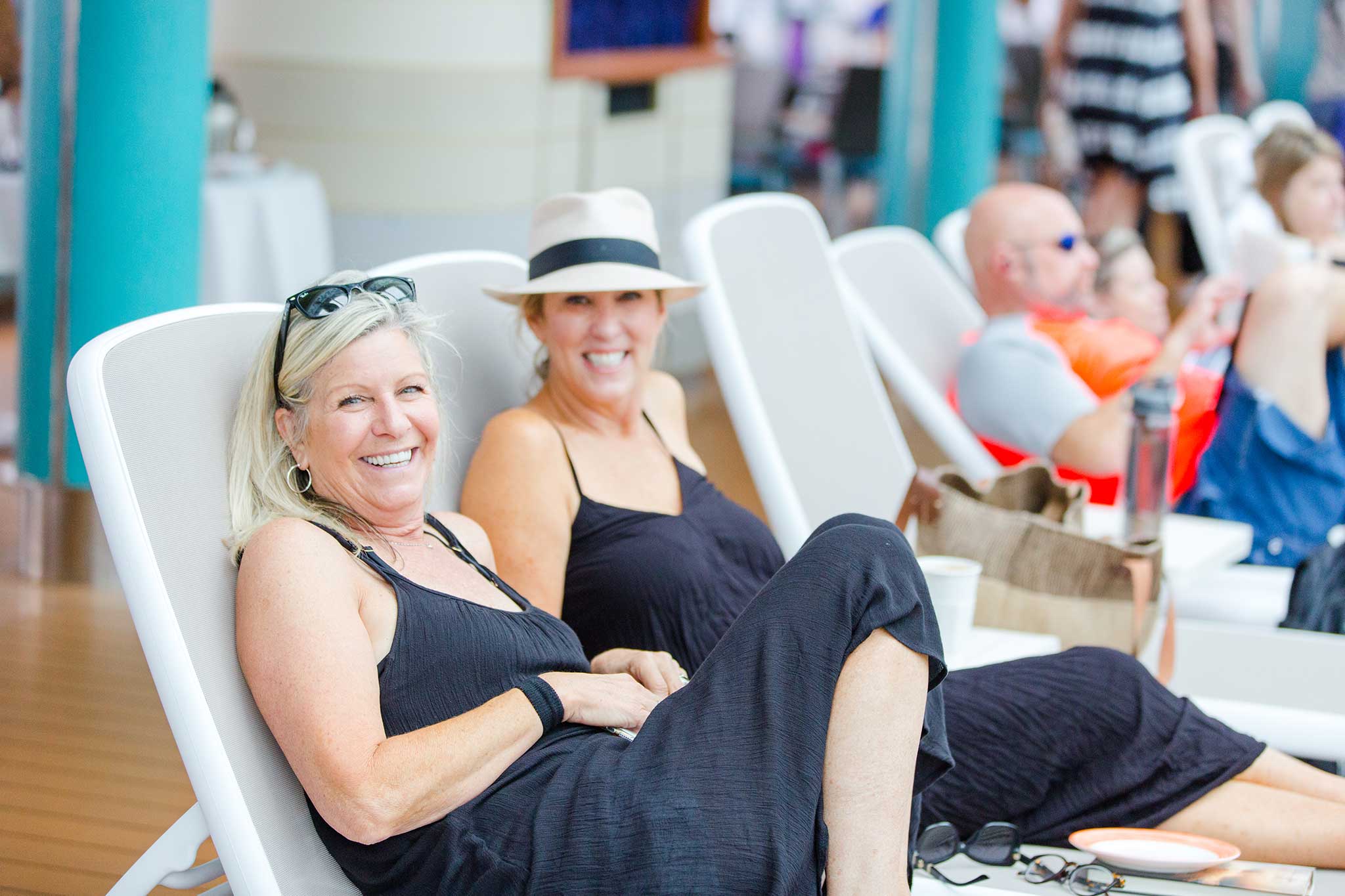 Two middle-aged ladies smile for the camera while wearing black swimsuit covers, hats and sunglasses while lounging on the cruise ship’s deck chairs.