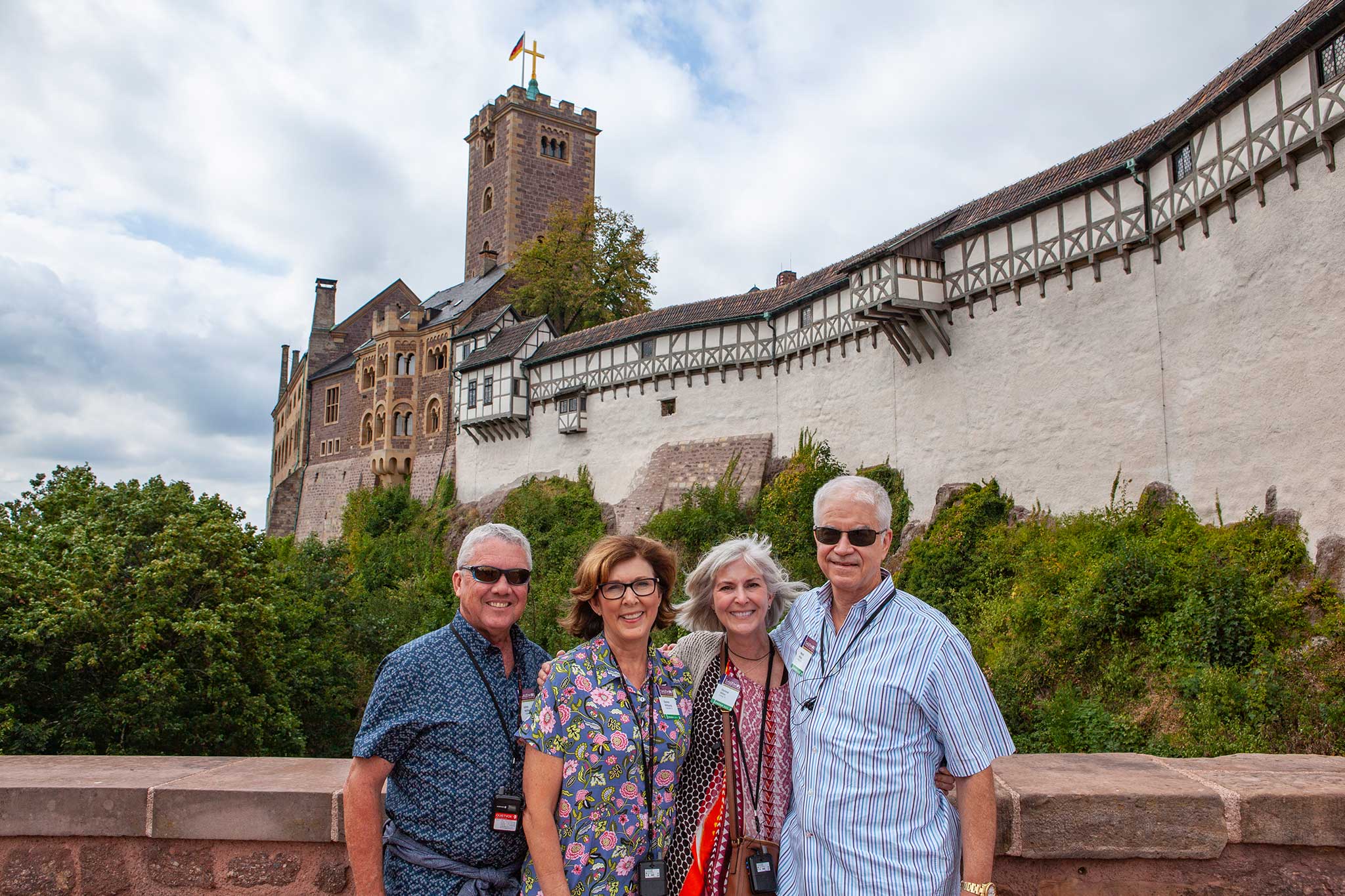 Two couples in different bright patterned shirts smile for the camera in front of green shrubbery and a brown brick castle towering in the background.