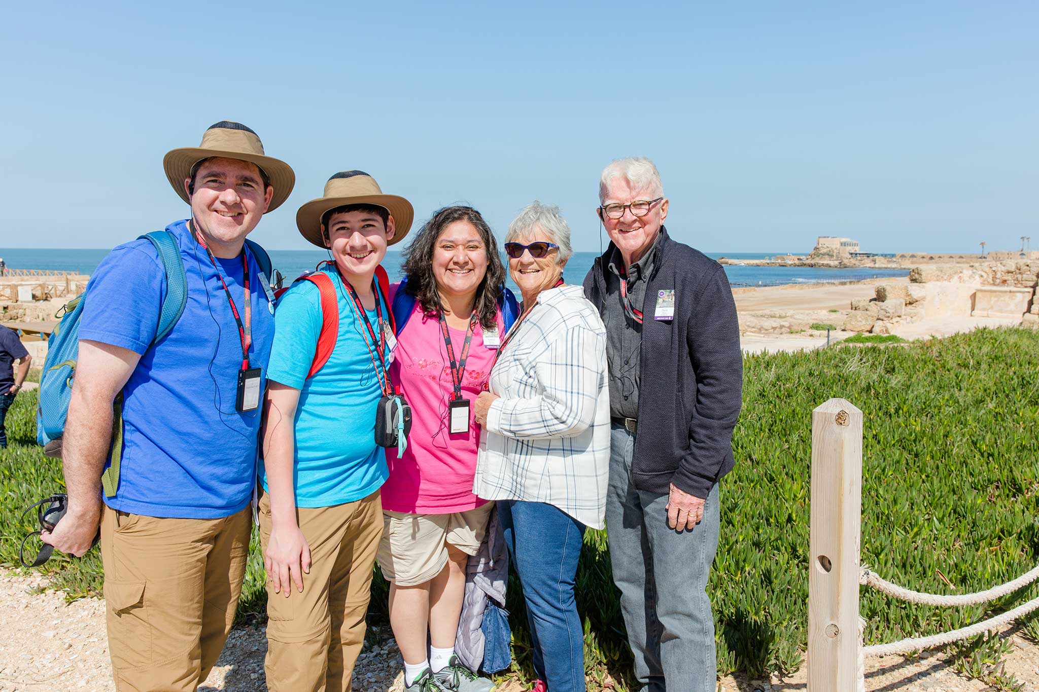 A multi-generational family smiles for the camera on a sunny day in front of coastal plants and a rocky tan beach along the Mediterranean Sea.