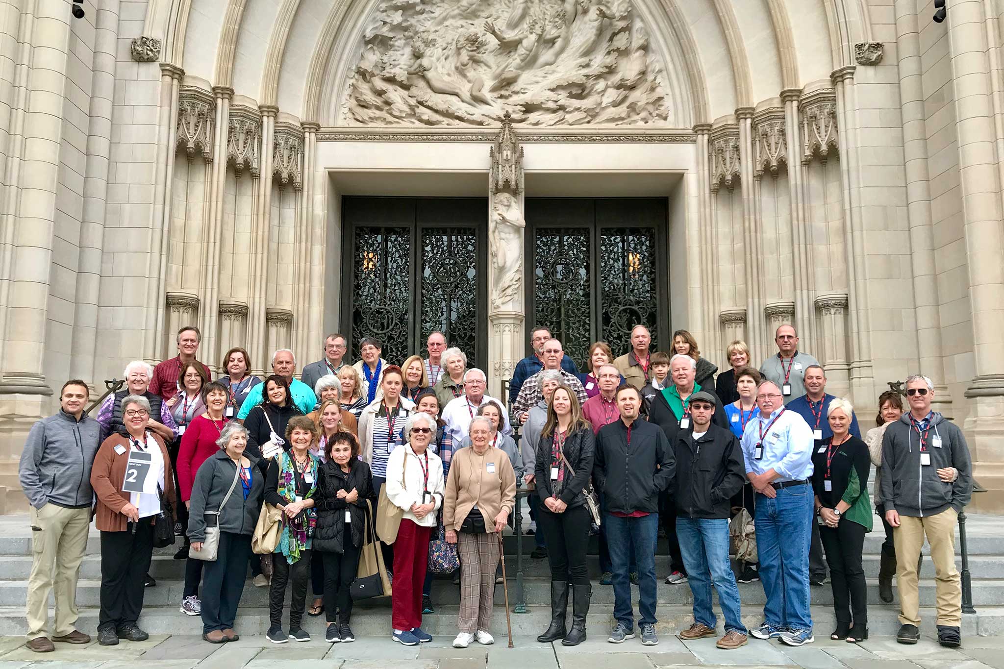 A group of older travelers stand together smiling for the camera in front of the entrance to the National Cathedral in Washington, D.C.