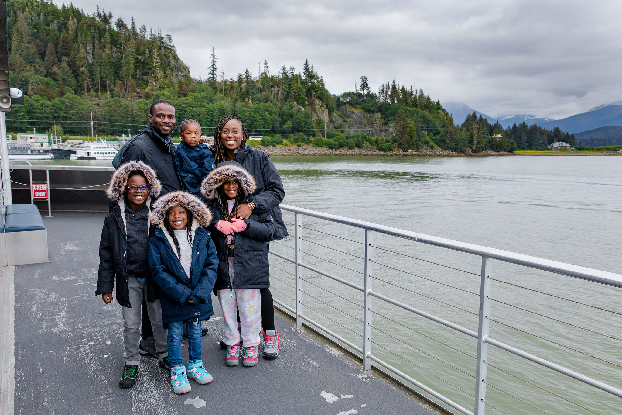 A family of six stands on a ferry deck, warmly dressed in winter coats, with Alaska’s gey waters and green, rugged hills in the background.