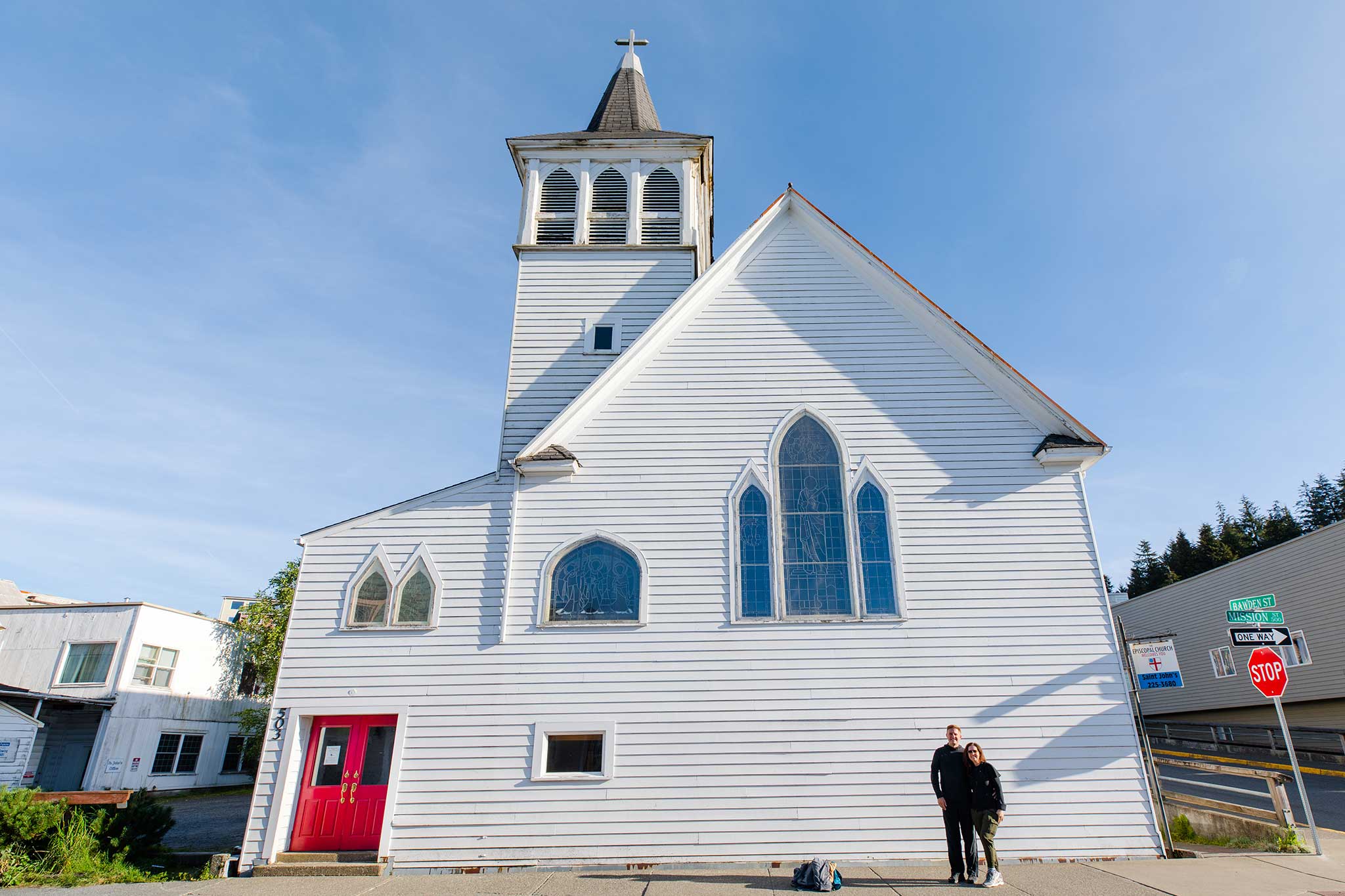 Classic white church with stained glass windows and a couple posing in front on a sunny day.