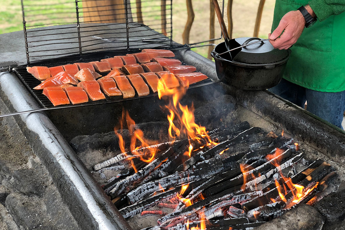 A cook prepares salmon on a barbecue with blazing coals.