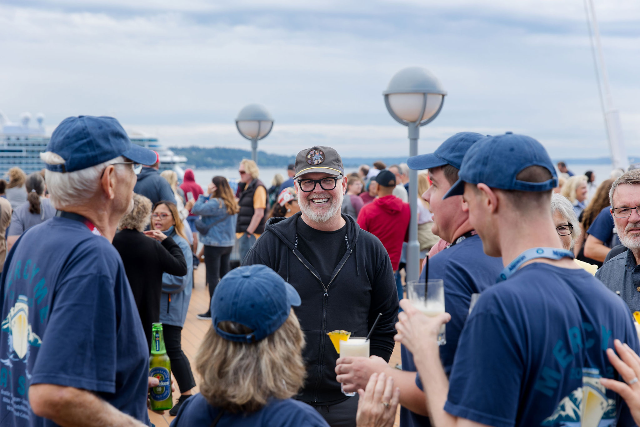 A smiling man in a black hoodie and cap chats with a group of people in blue shirts during a cruise deck gathering.