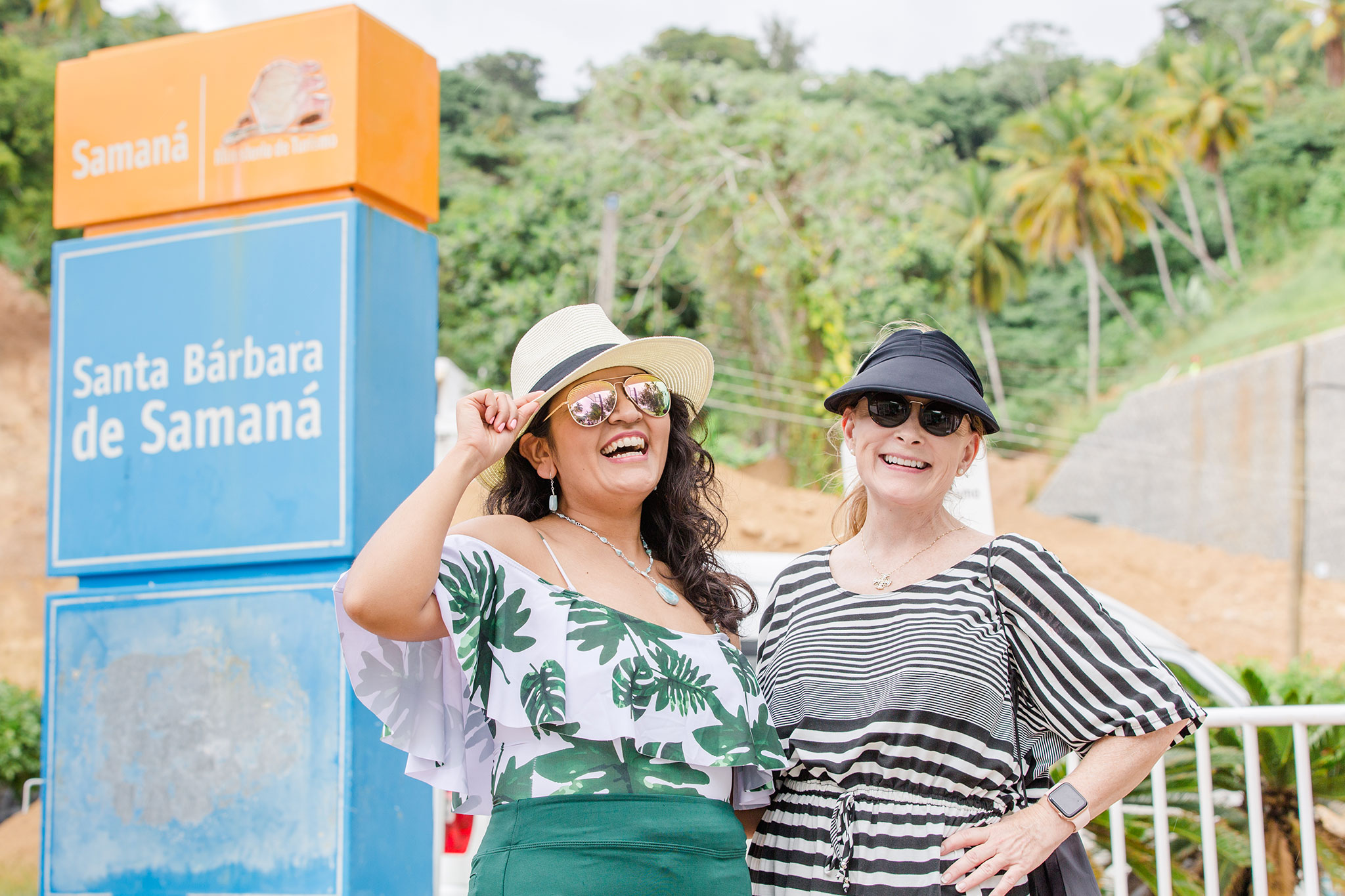 Two women smiling and posing together in sun hats and sunglasses beside a blue and orange Santa Barbara de Samana sign, with palm trees and lush tropical greenery behind them.