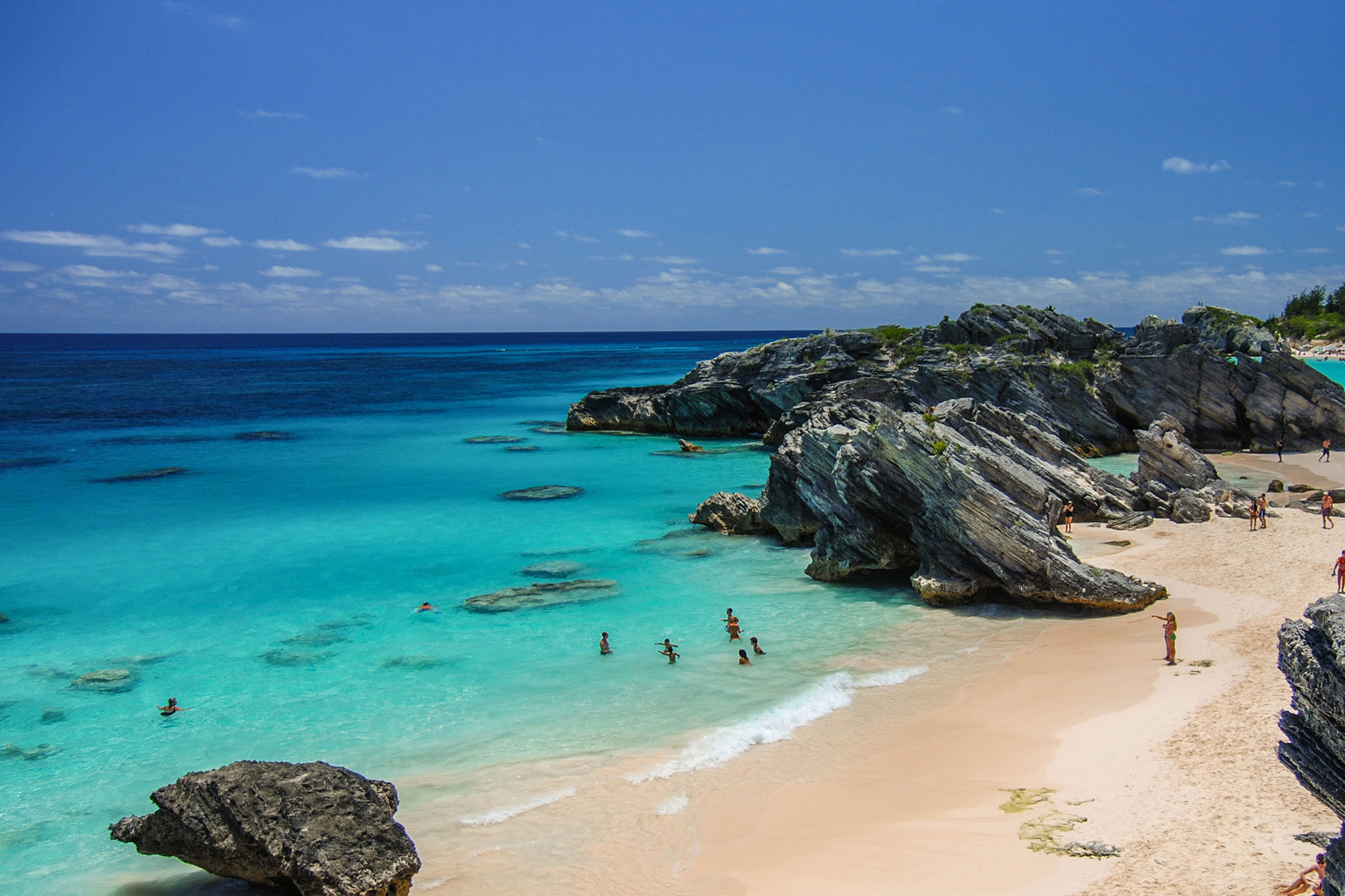 Turquoise ocean water laps against a soft sandy beach framed by rugged limestone rock formations, with people swimming, walking along the shore and relaxing under a bright blue sky.
