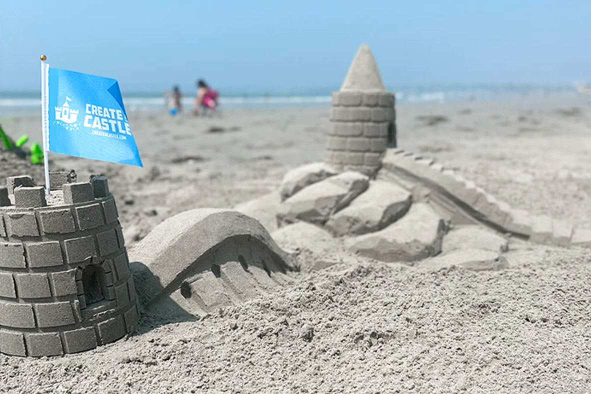 Sandcastle with blue Create A Castle flag on a beach.
