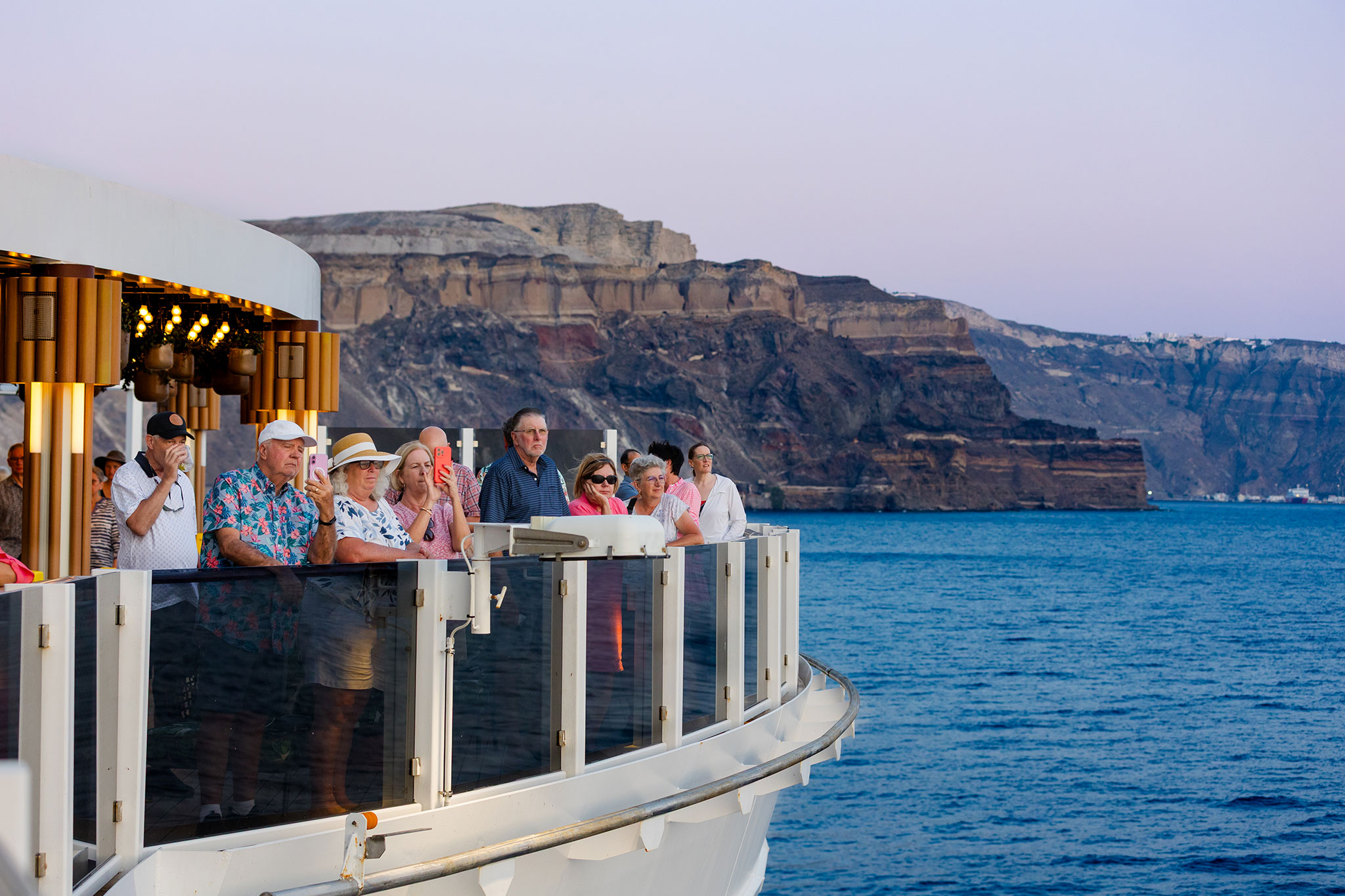 Travelers line the railing of a cruise ship’s upper deck, looking out over deep blue water as the ship sails past tall, rugged coastal cliffs in the evening light.