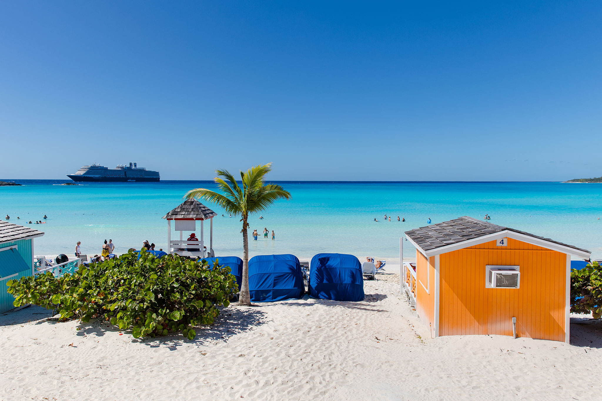 A sunny beach scene with white sand, turquoise ocean water, and people wading and relaxing near shore, framed by colorful beach huts, blue loungers, and a palm tree. A cruise ship is anchored in the distance under a clear blue sky.