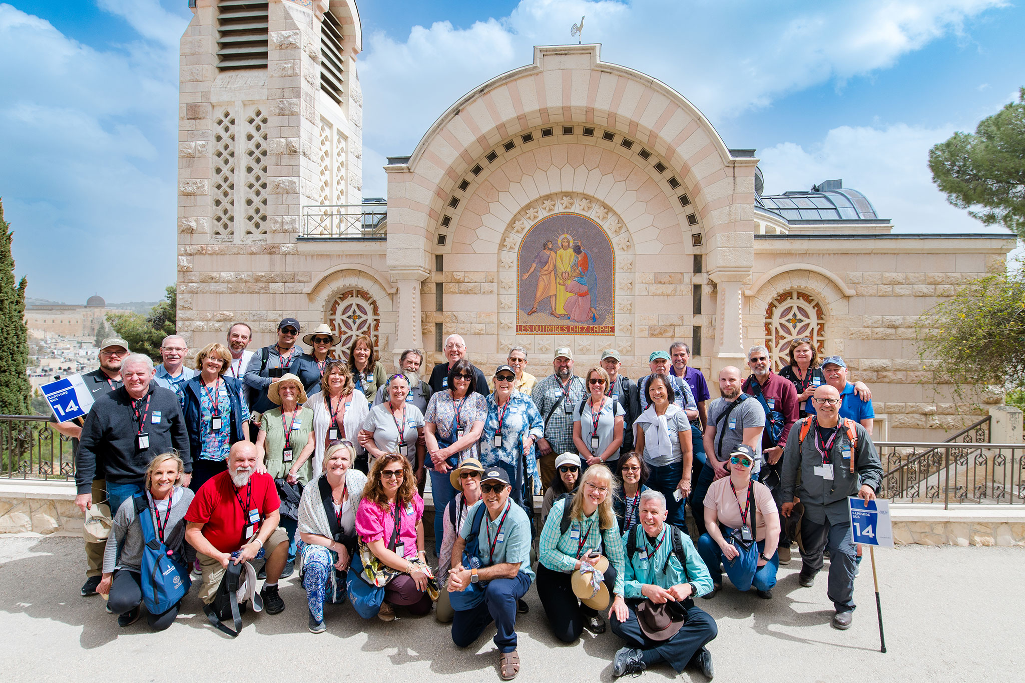 A large group of travelers wearing lanyards and backpacks poses together in front of the House of Caiaphas, a stone church with an arched facade and colorful mosaic, beneath a bright blue sky in Jerusalem.