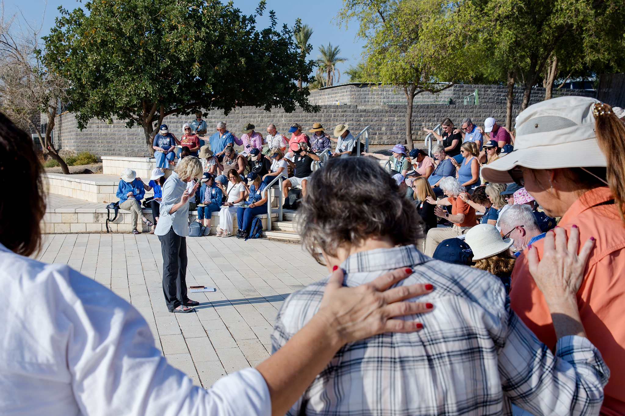 Travelers seated on stone steps beneath trees gather in prayer at Capernaum.  A group leader speaks while participants bow their heads and rest their hands on one another’s shoulders in the sunlit courtyard.