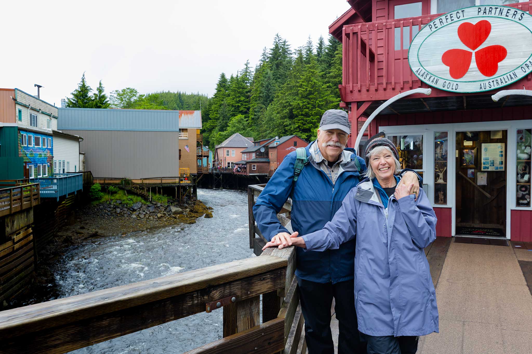 A smiling couple stands on a wooden boardwalk along Creek Street in Ketchikan, Alaska, with colorful buildings on stilts over the creek, a local shop entrance, and lush forested hills in the background.