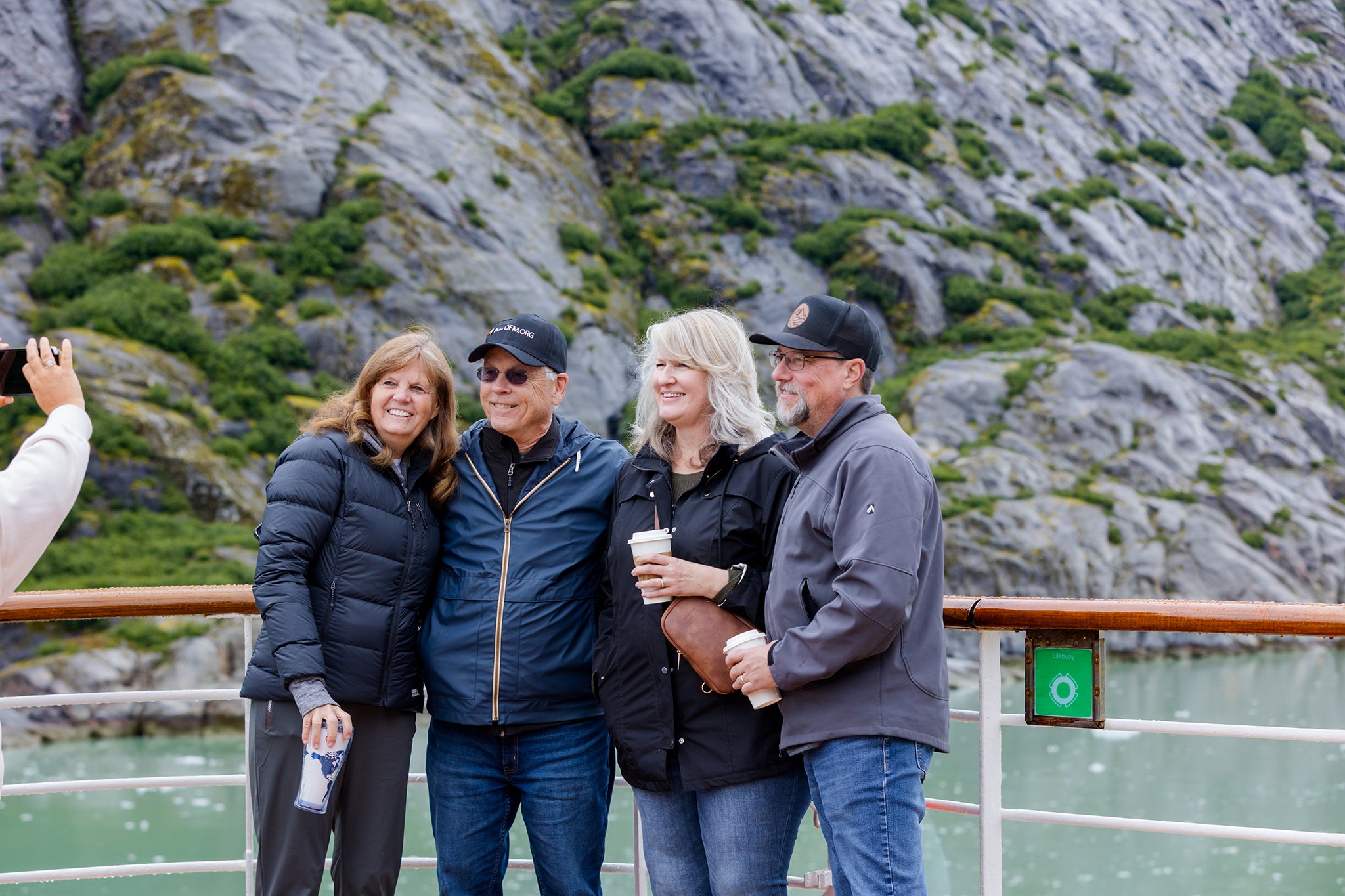 Four travelers stand together on a cruise ship deck on an Alaska Christian cruise, smiling for a photo as one person off to the side takes their picture. Behind them, steep, rocky cliffs and calm water create a scenic backdrop as the group holds warm drinks and leans in close, enjoying the moment together.