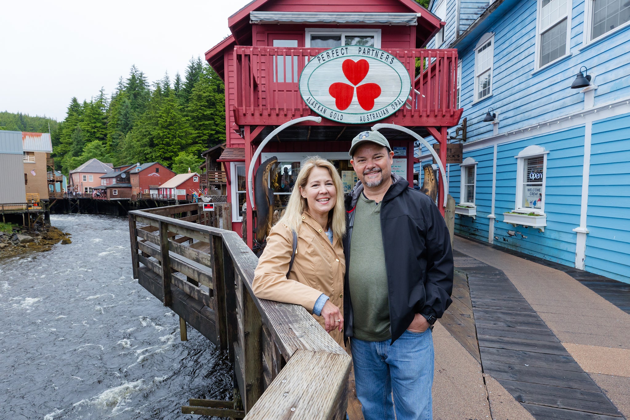 A couple stands together on a wooden boardwalk beside a rushing creek, smiling for a photo in a colorful coastal town. Behind them, a red building with a “Perfect Partners” sign and nearby pastel storefronts line the waterfront, surrounded by lush green trees.