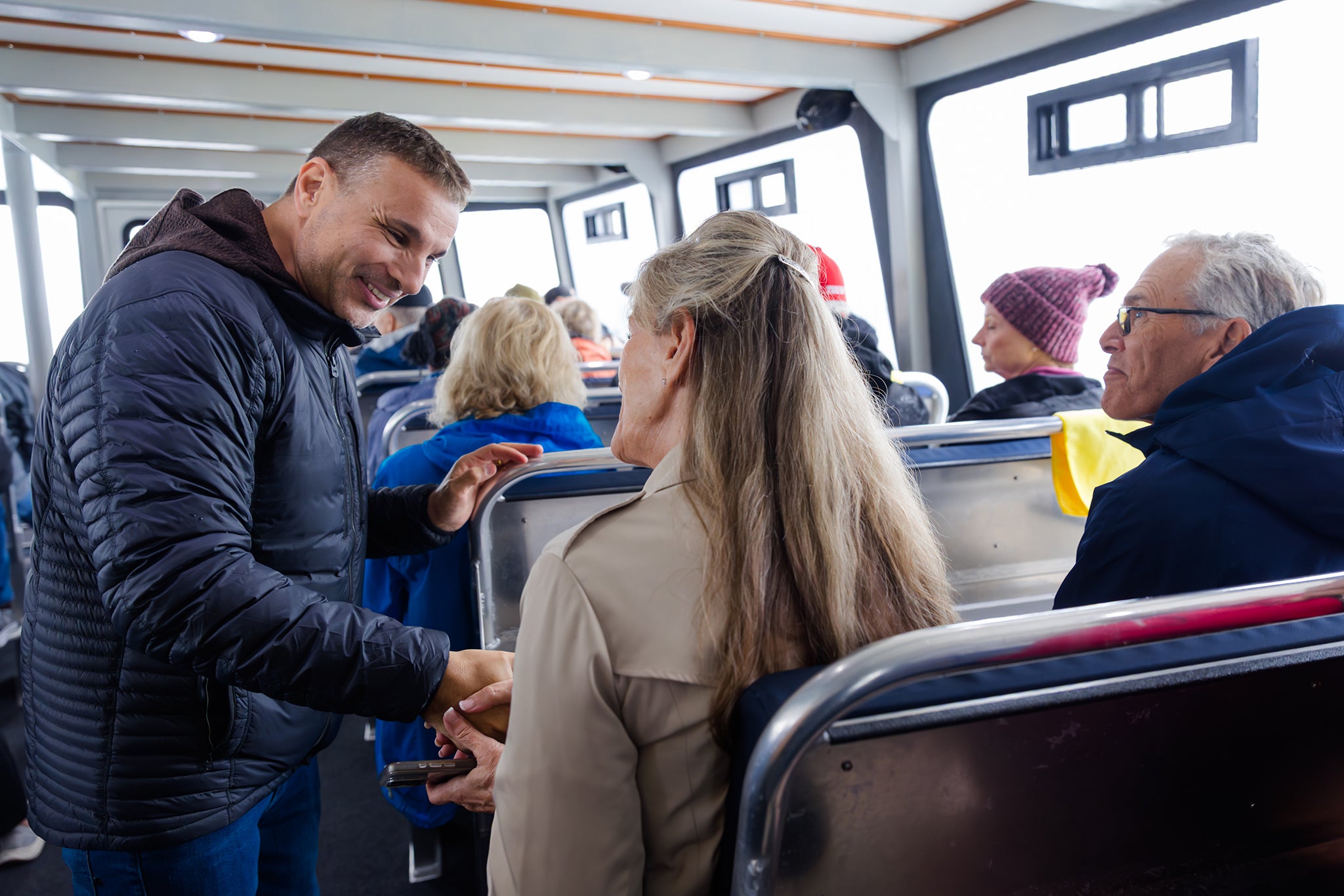 Amir Tsarfati smiles and greets fellow travelers on a passenger ferry, shaking hands and speaking with a seated woman as others look on in the bright cabin.
