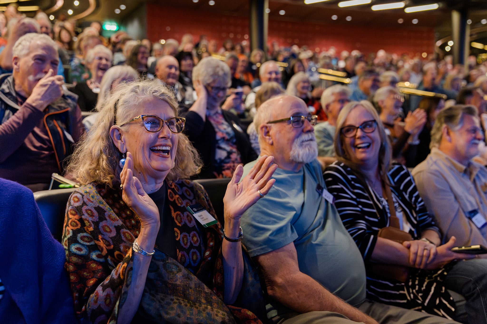A packed theater aboard a cruise ship is filled with smiling travelers, many laughing and applauding as they enjoy a live event. In the foreground, a woman with glasses claps enthusiastically while others around her grin and react, capturing the joyful, shared energy of the onboard experience.