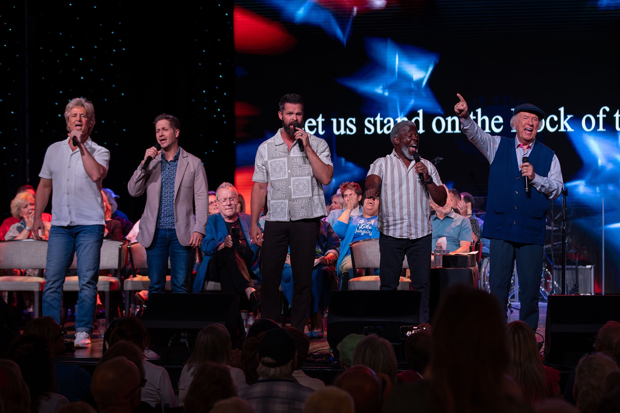 Five male vocalists stand across a stage singing into microphones during a live concert. A seated choir is visible behind them and a large screen displays blurred red, white and blue stars with partial text.