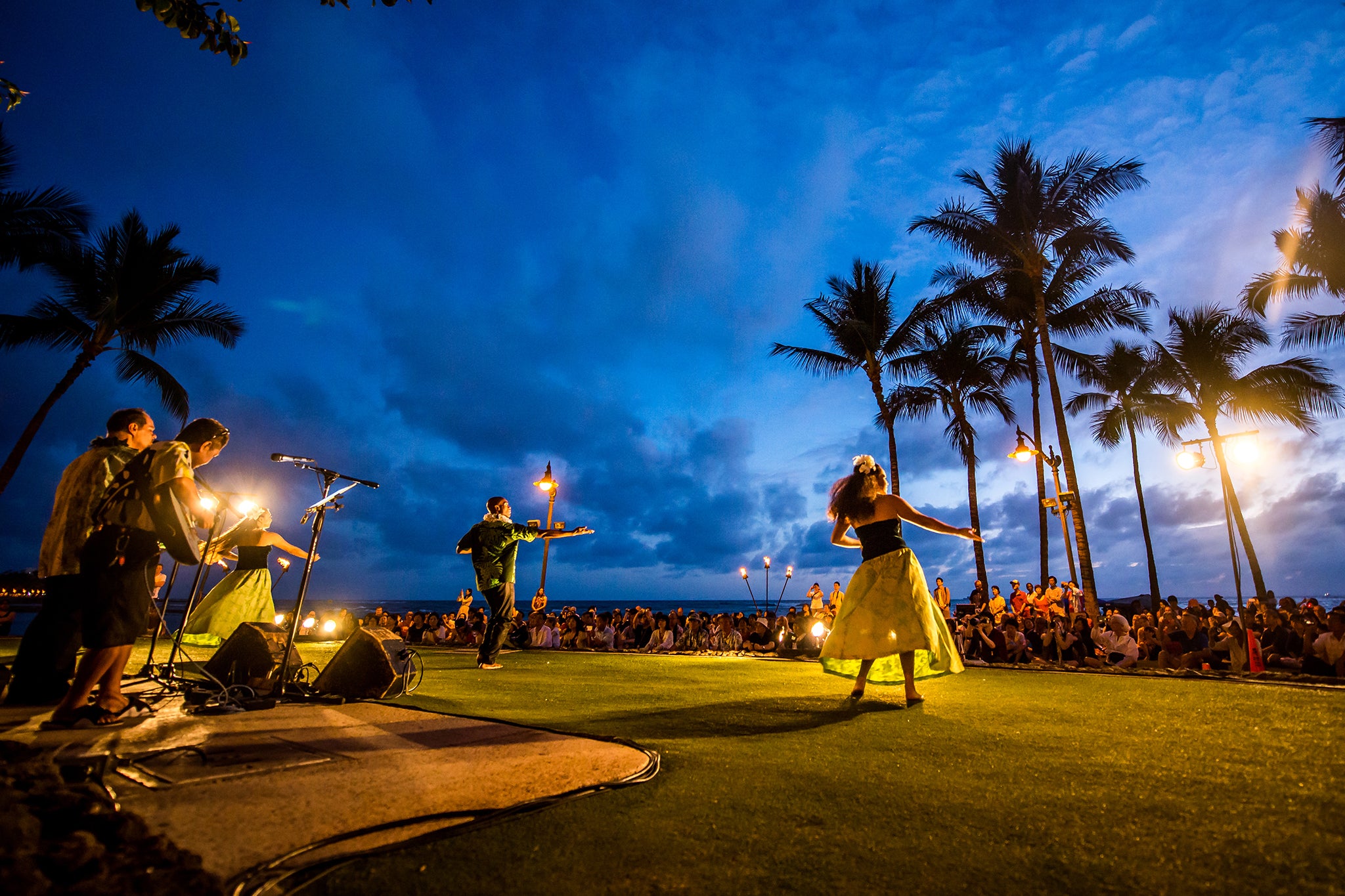 Outdoor evening performance on a tropical beachfront with palm trees silhouetted against a deep blue sky, as musicians and hula dancers perform on a lit stage before a seated crowd gathered on the lawn.