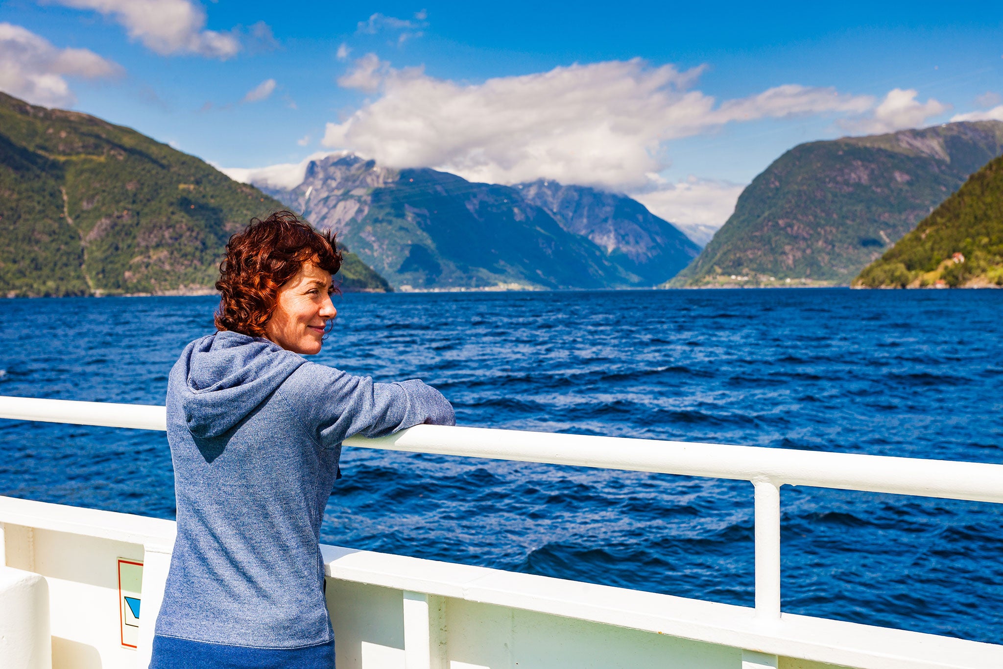 Traveler standing on the deck of a ferry or cruise ship, looking out across a deep blue Norwegian fjord surrounded by towering green mountains and distant snow-capped peaks beneath a bright sky with scattered clouds.