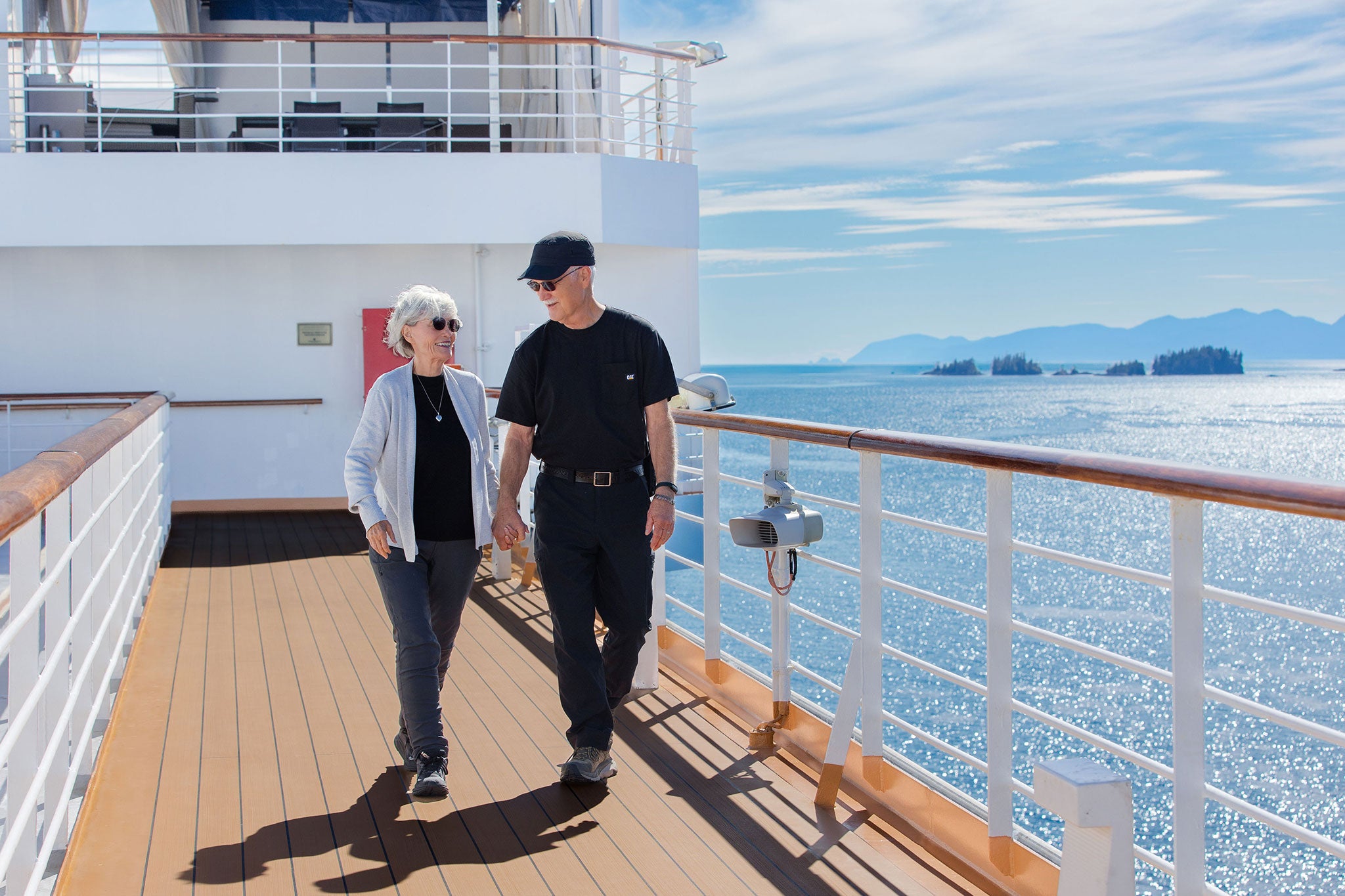 A smiling older couple walking hand in hand along a cruise ship deck on a sunny day. Sparkling blue water, distant forested islands and mountain silhouettes create a scenic backdrop as they enjoy time together at sea.