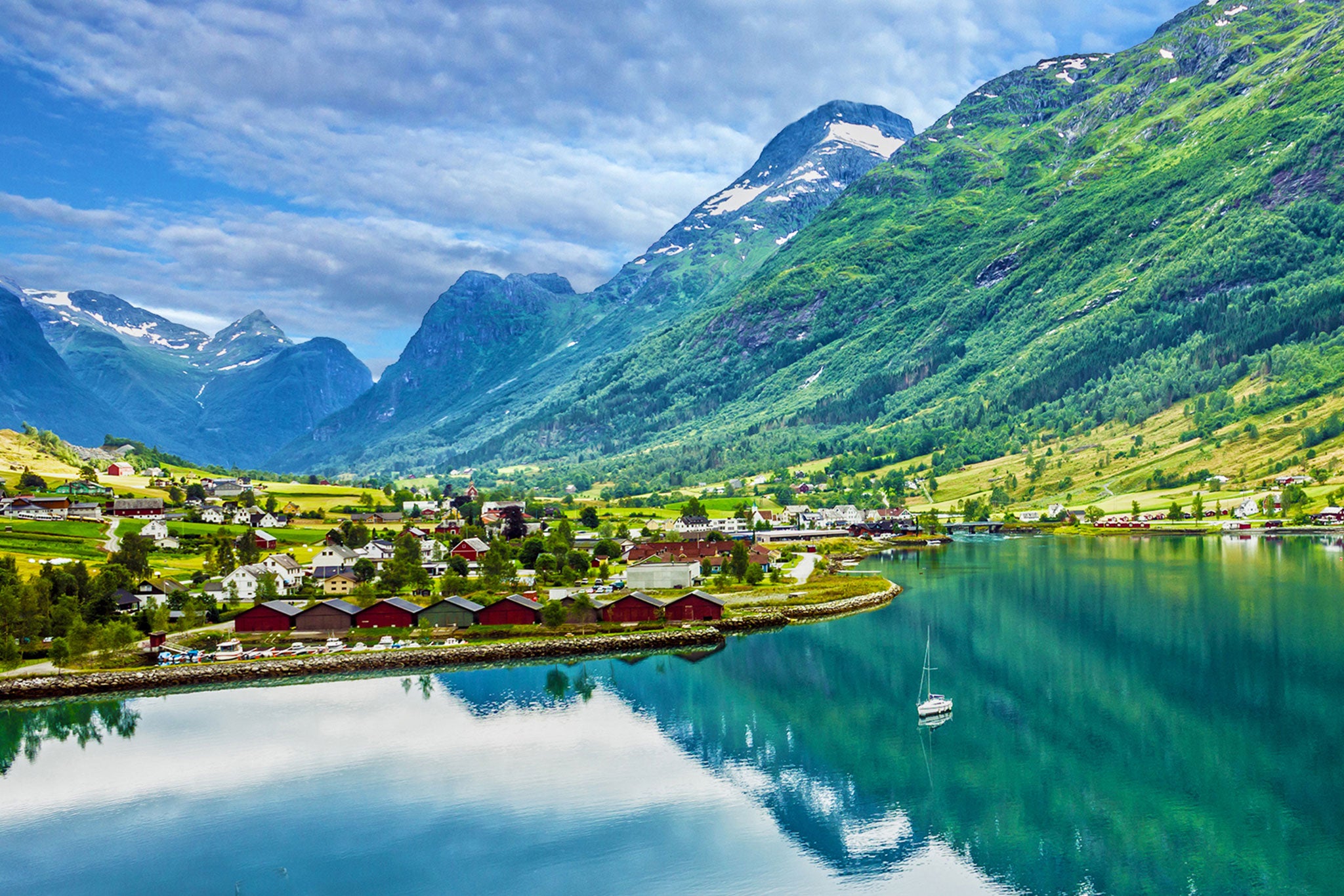 Panoramic view of a Norwegian fjord surrounded by towering green mountains and a picturesque village along the shoreline. Calm turquoise water reflects the dramatic landscape, while a lone sailboat rests in the center of the fjord beneath a cloud-filled sky.