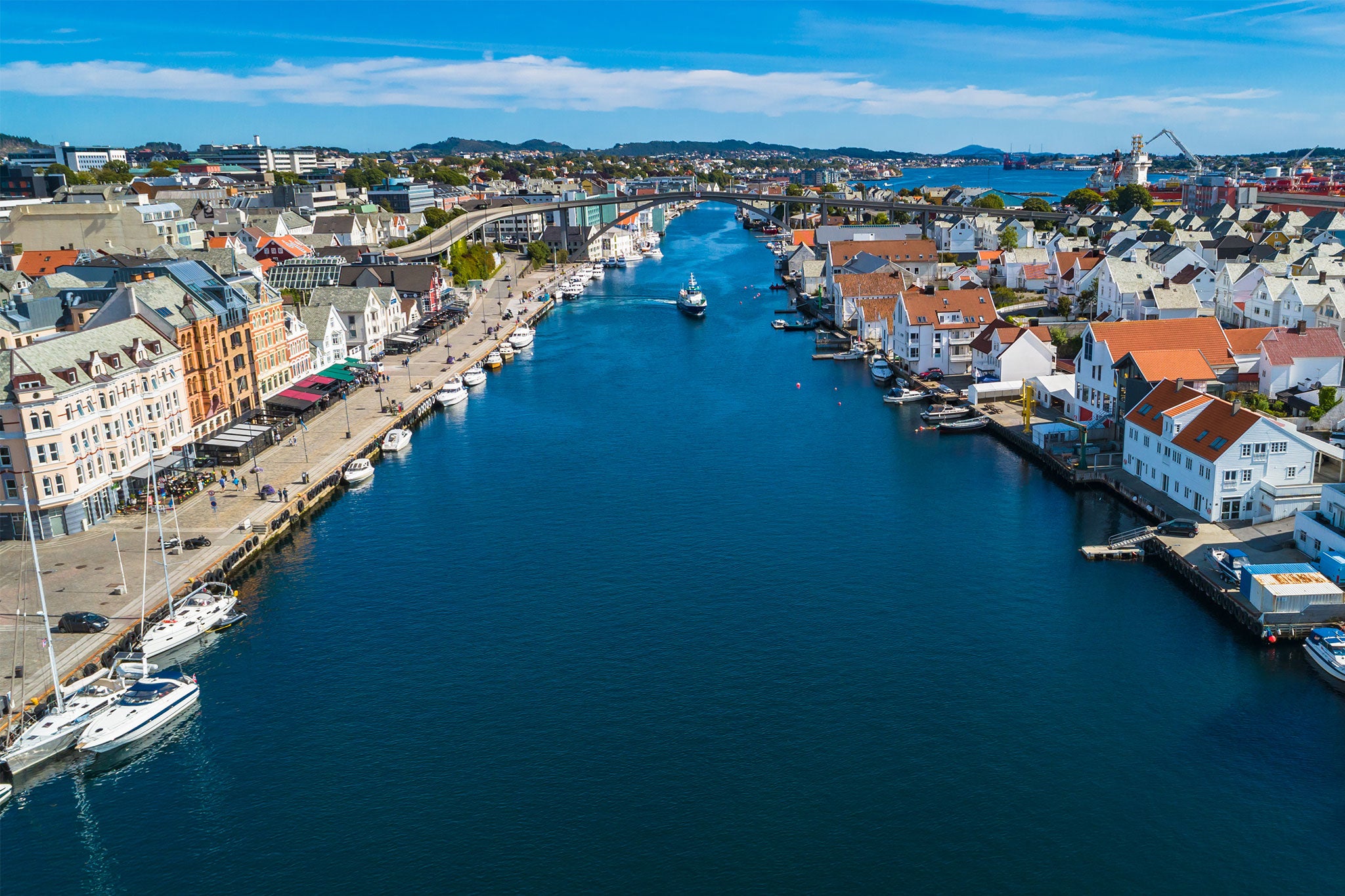 Aerial view of a coastal Norwegian town with colorful waterfront buildings lining a narrow harbor channel. Boats dot the deep blue water, while bridges, shipyards and clusters of homes stretch across the shoreline beneath a clear sky.