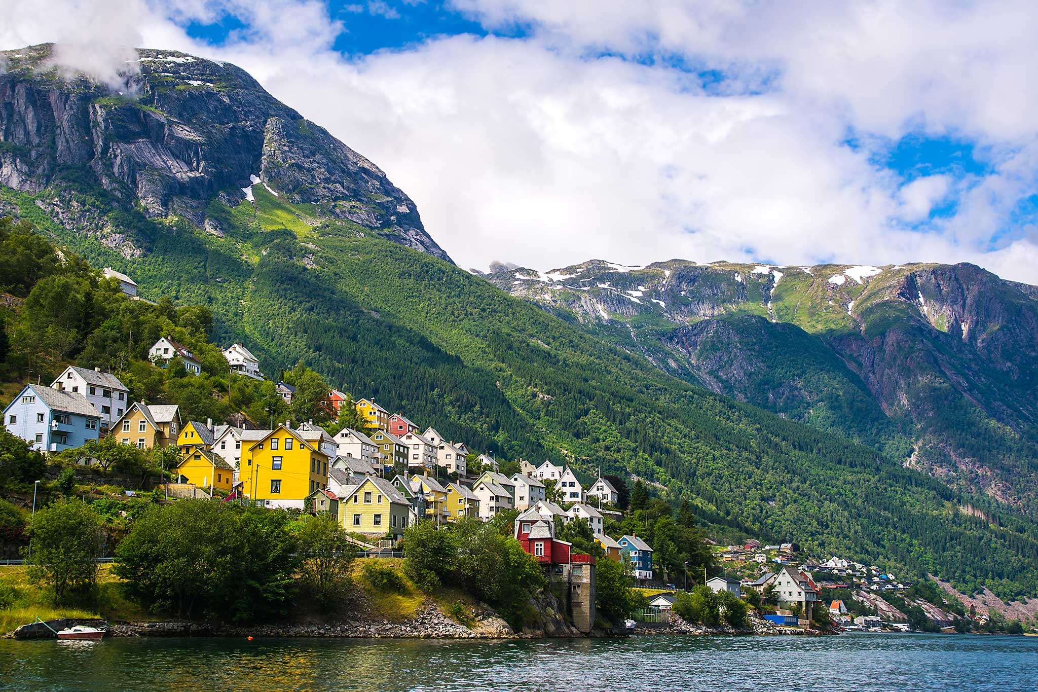 Colorful houses dot the shoreline of a Norwegian fjord beneath towering mountains covered in lush forests and patches of snow. The peaceful waterfront village sits at the base of dramatic peaks under a bright sky filled with scattered clouds.