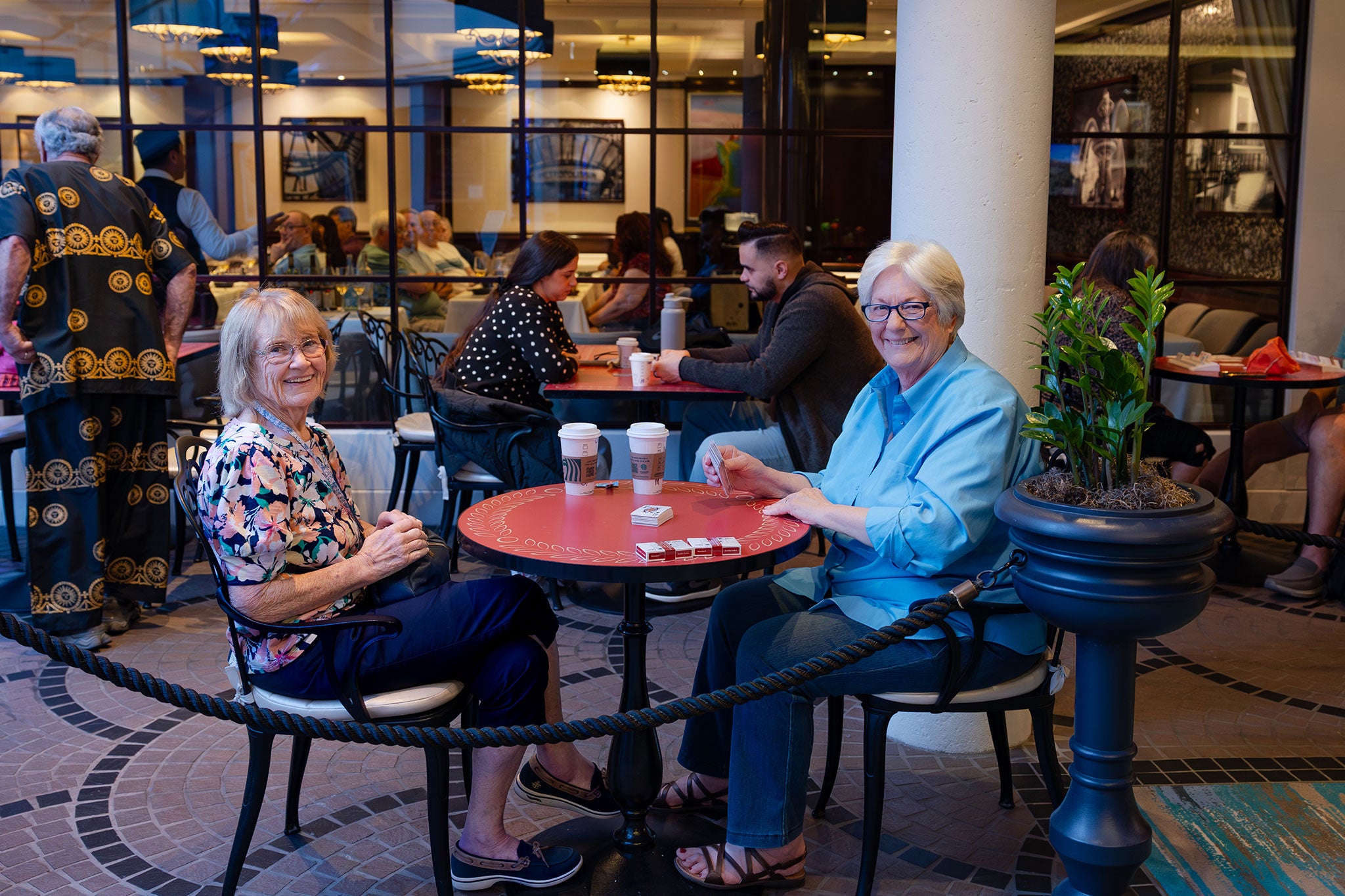 Two women smile while playing cards at a small cafe table aboard a cruise ship. Coffee cups sit nearby as fellow travelers gather and socialize in the warmly lit lounge area around them.