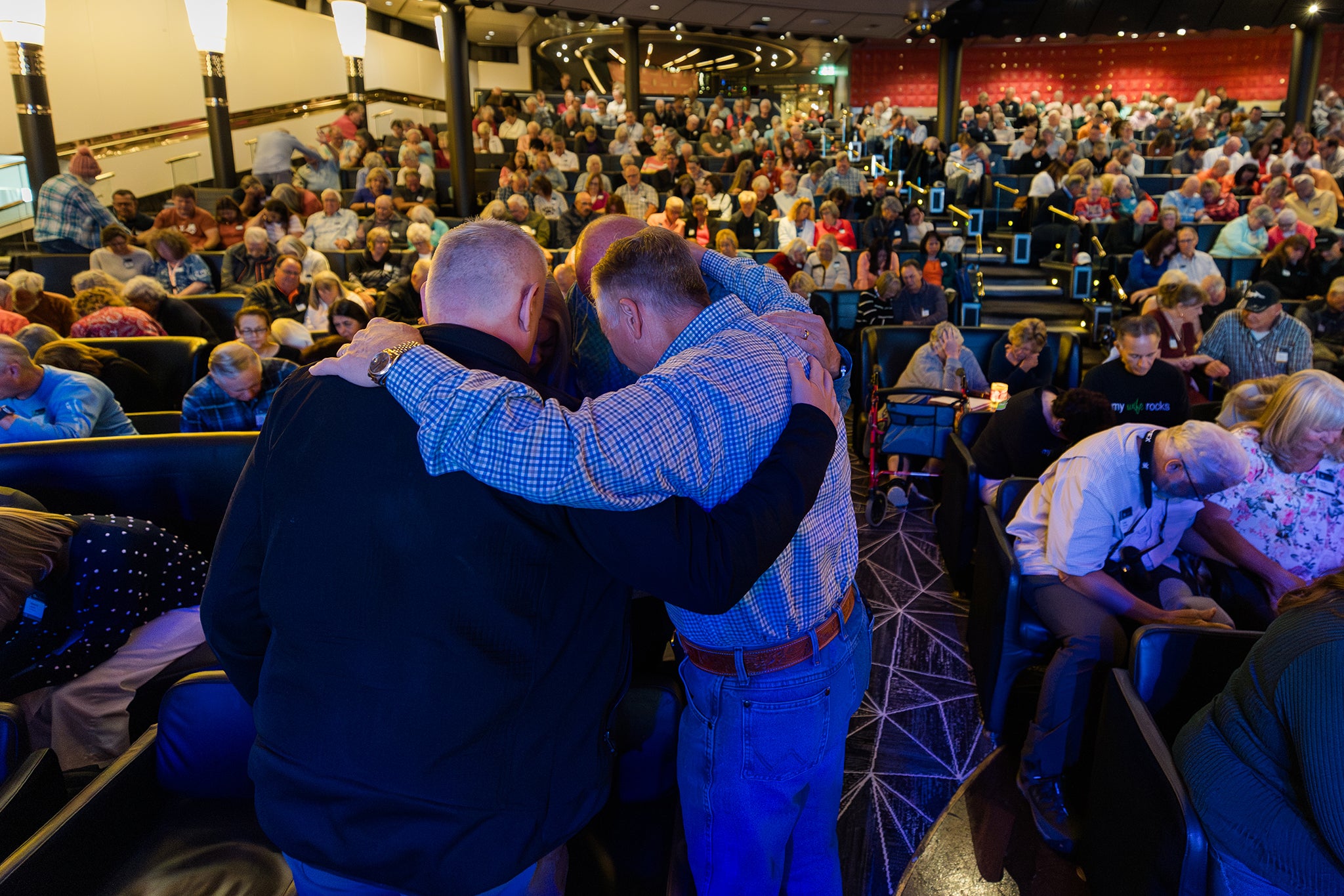 Travelers fill a cruise ship theater as many bow their heads in prayer during a worship gathering. In the foreground, several attendees stand together with arms around one another in a moment of fellowship and encouragement.