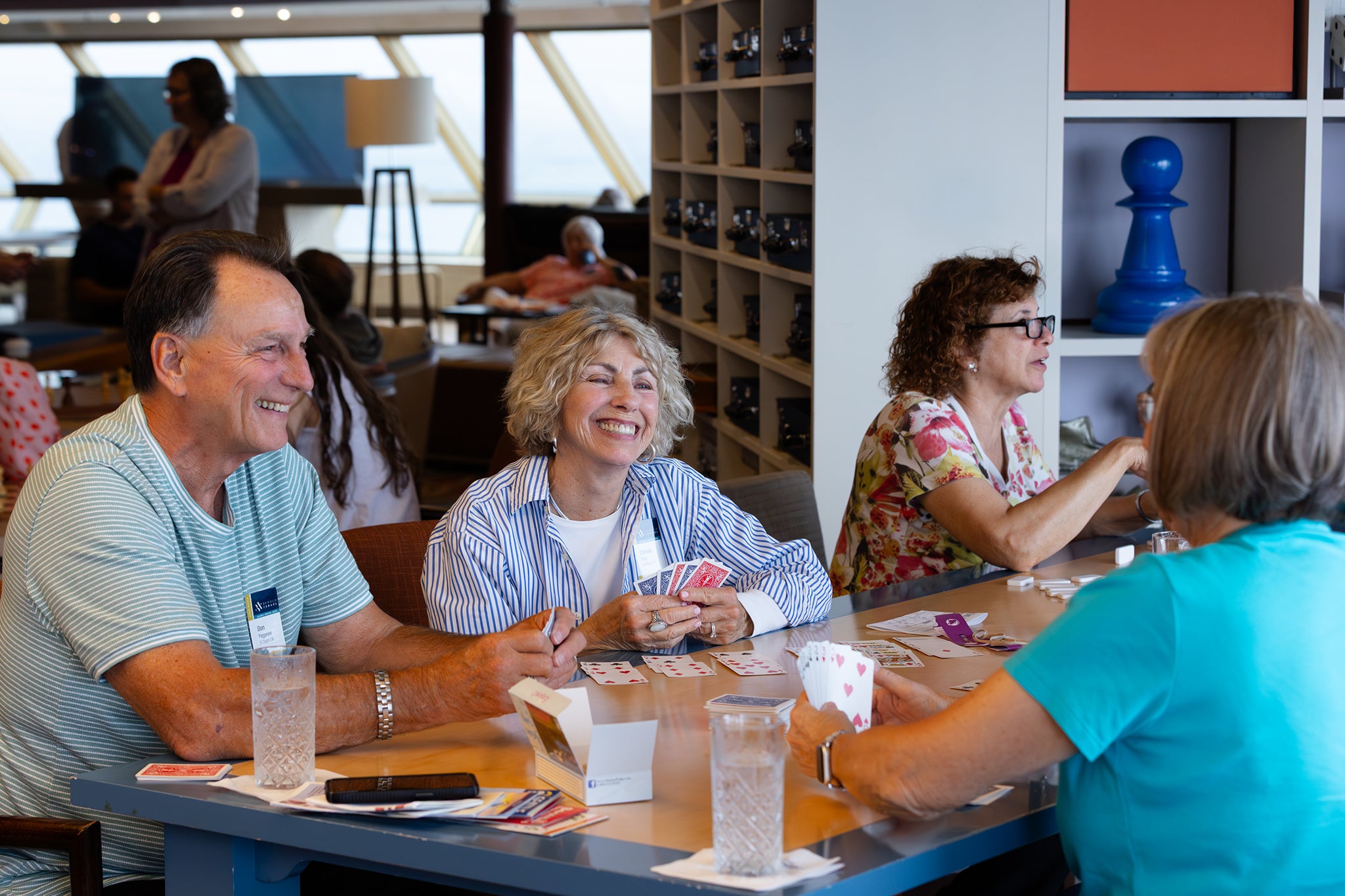 Several travelers gather around a table aboard a cruise ship, smiling and playing cards together. The relaxed setting highlights the fellowship and friendships that develop while traveling.