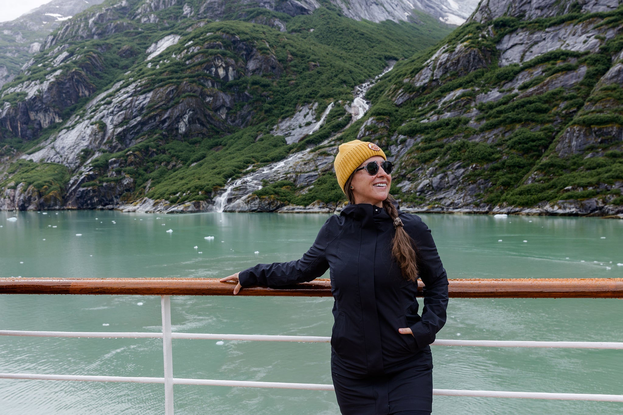 A traveler stands on the deck of a cruise ship, taking in views of a glacier-carved fjord surrounded by rugged mountains and cascading waterfalls. The serene landscape showcases the breathtaking beauty of Alaska’s wilderness.