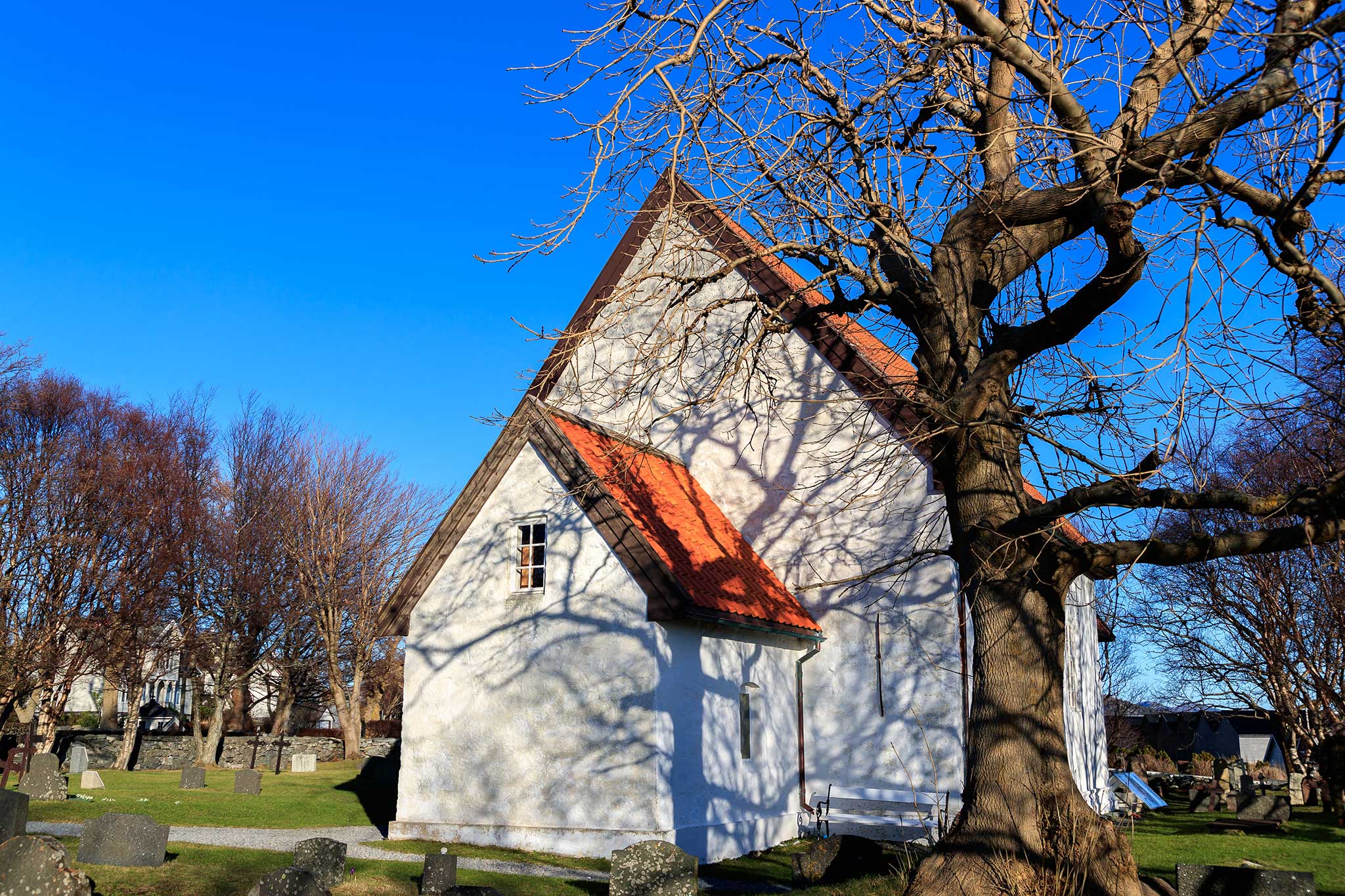 A small white church with a red roof stands beside a large leafless tree, surrounded by gravestones and a grassy churchyard. Bright blue skies and bare trees frame the peaceful historic scene.