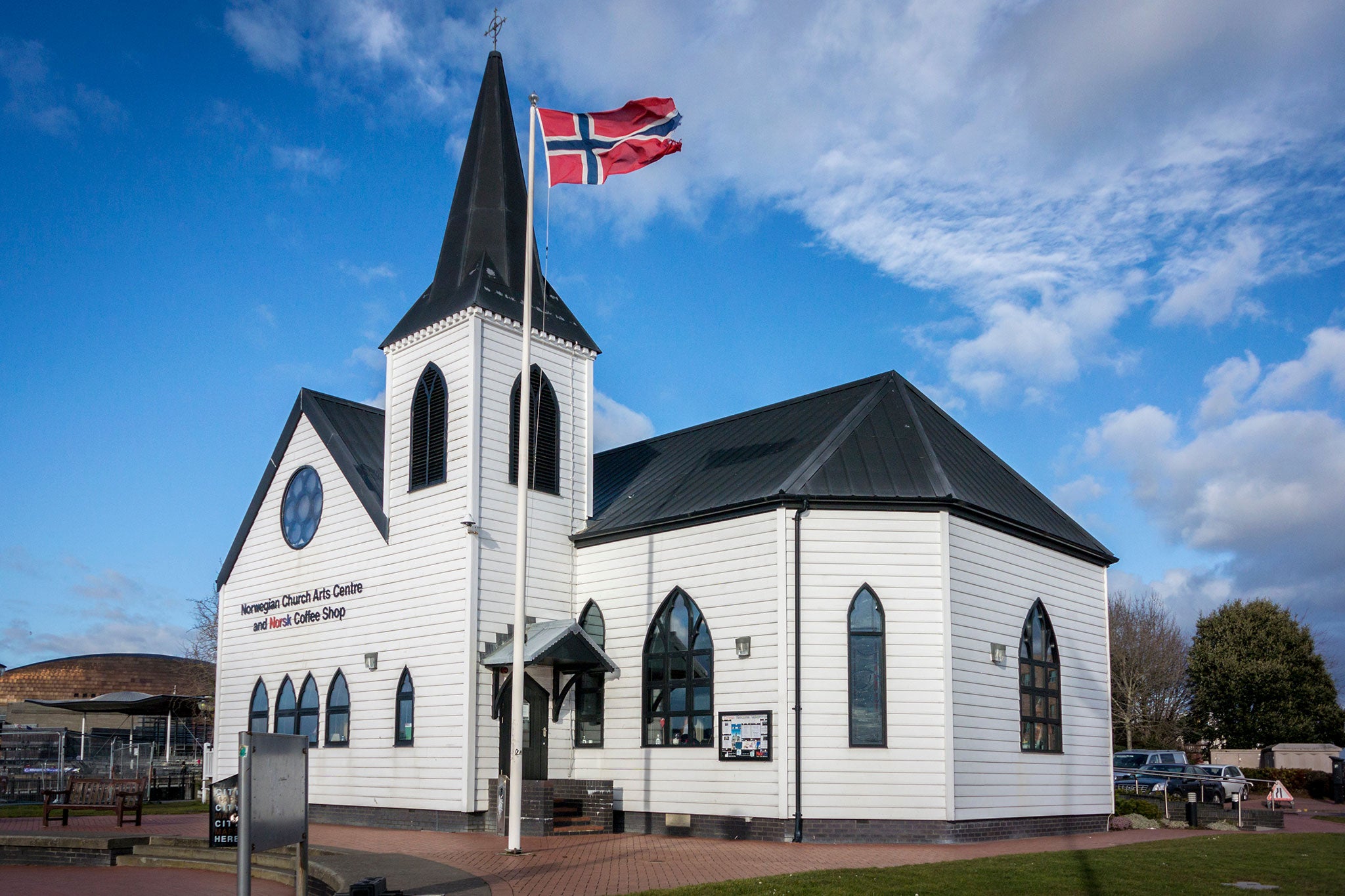 A white church with a black steeple stands beneath a bright blue sky, with a Norwegian flag flying in front. The church’s traditional architecture and waterfront setting reflect Norway’s rich cultural heritage.