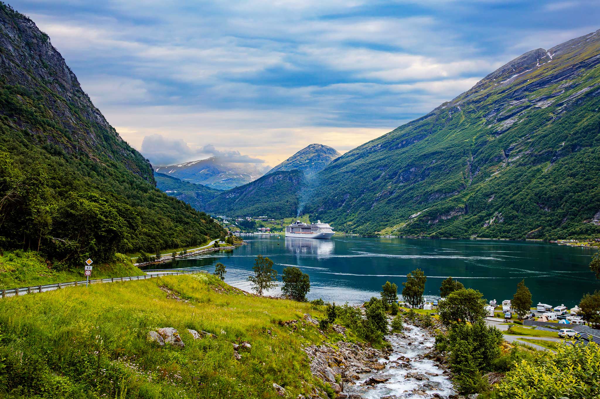 A cruise ship sails through a tranquil Norwegian fjord surrounded by towering green mountains and cascading streams. The dramatic landscape showcases the breathtaking beauty of Norway’s rugged coastline.