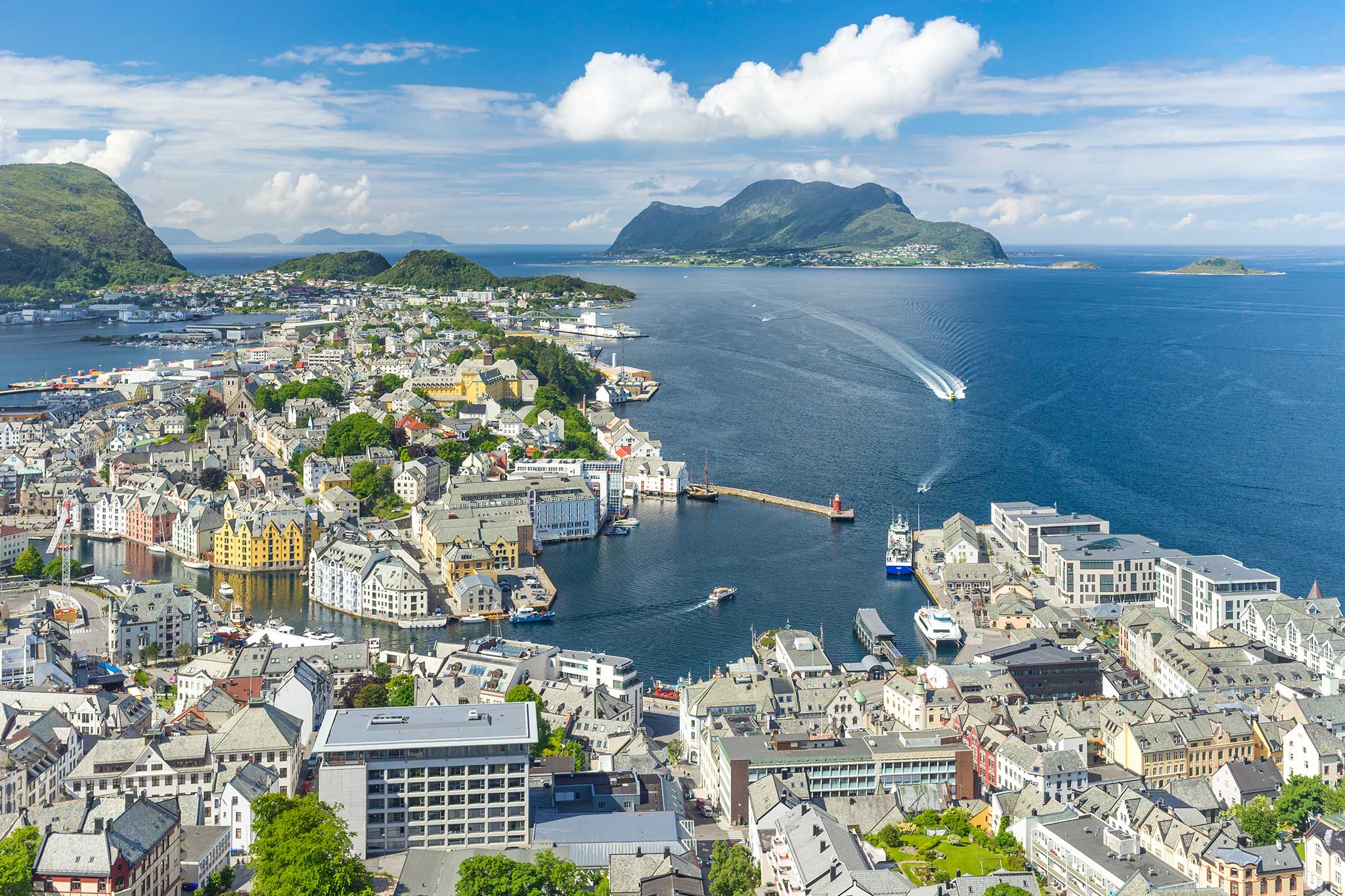 Aerial view of a coastal Norwegian city with colorful waterfront buildings, a busy harbor and surrounding islands. Blue waters and distant mountains create a stunning backdrop to the seaside landscape.