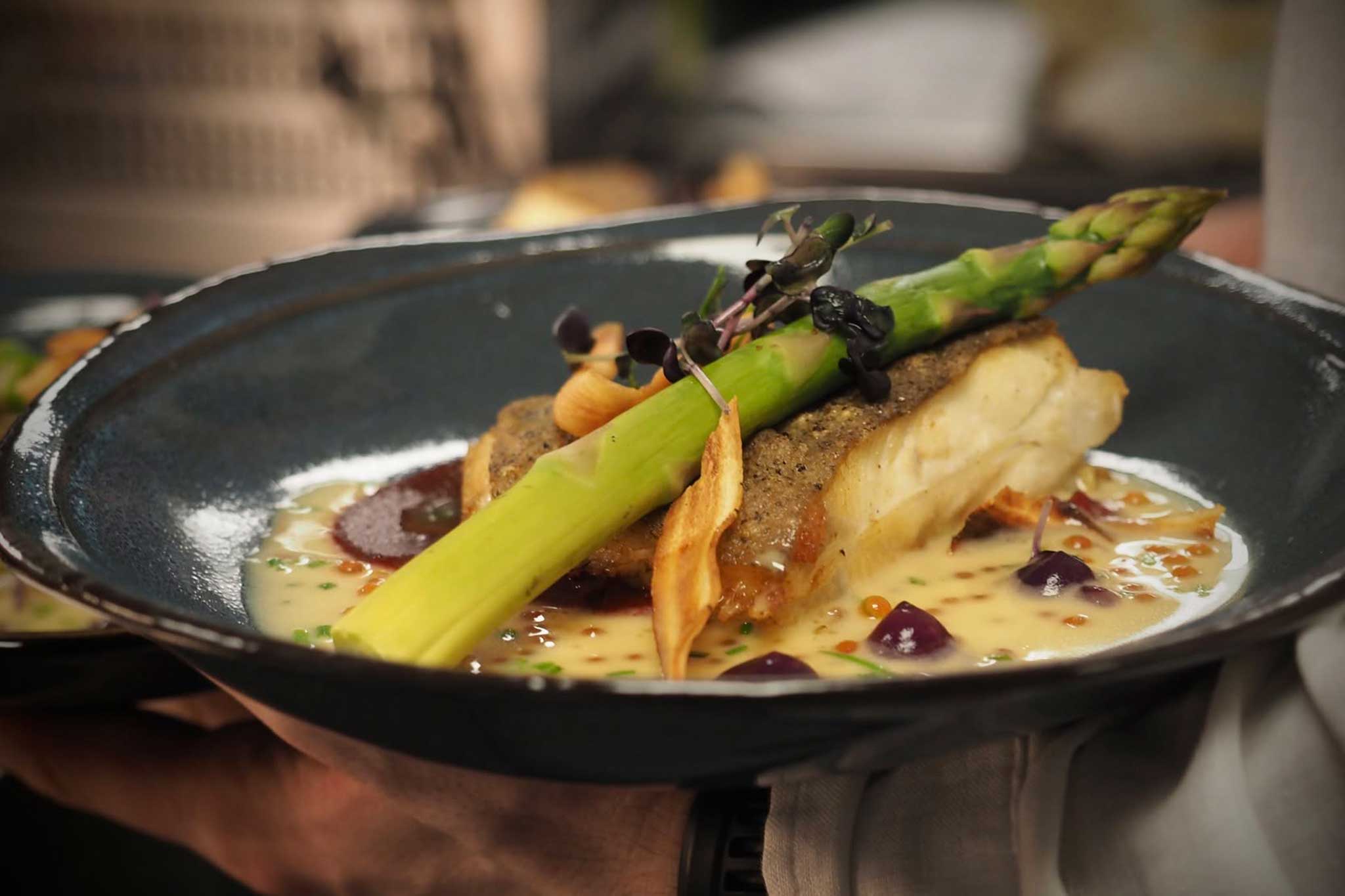 A plated gourmet dish featuring a fish fillet topped with fresh asparagus and microgreens, served in a creamy sauce. The elegant presentation highlights the fine dining experience enjoyed on board.