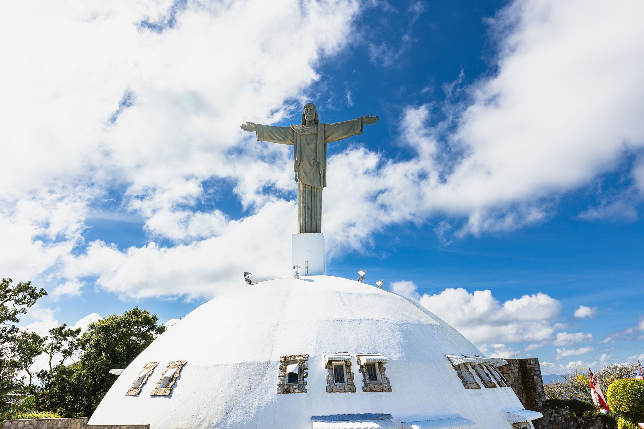 A towering statue of Christ with outstretched arms stands atop a large white domed structure beneath a vibrant blue sky filled with billowing clouds. Trees and stone architecture surround the hilltop landmark, creating a striking scene of faith and Caribbean beauty.