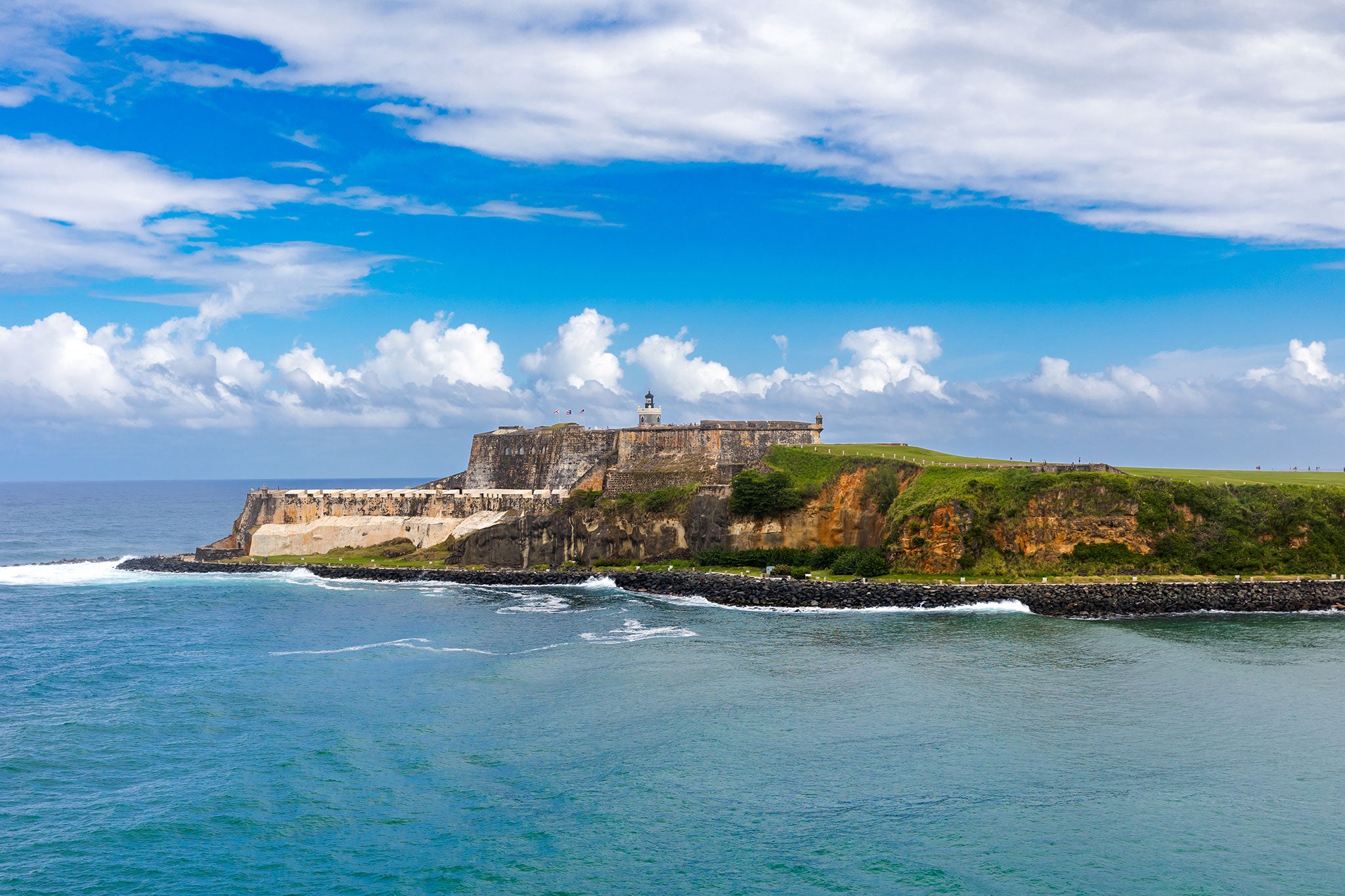 A historic stone fortress sits atop a rocky peninsula overlooking the turquoise waters of the Atlantic Ocean. Surrounded by rugged cliffs and lush green terrain, the coastal landmark stands beneath a dramatic sky filled with billowing white clouds.