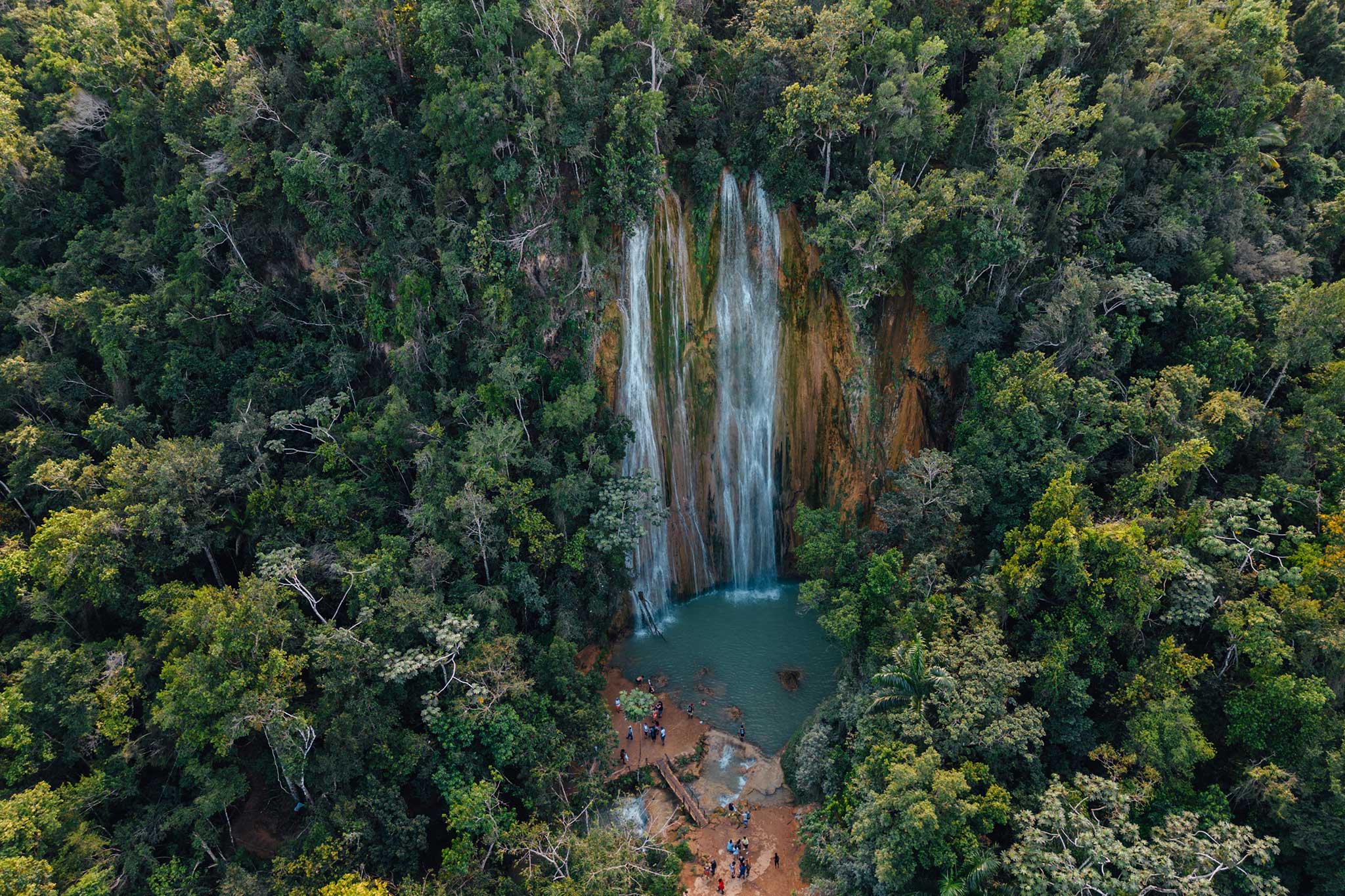 An aerial view of a tall waterfall cascading into a secluded pool deep within a dense tropical rainforest. Visitors gather at the base of the falls, surrounded by lush green trees and rugged natural terrain.