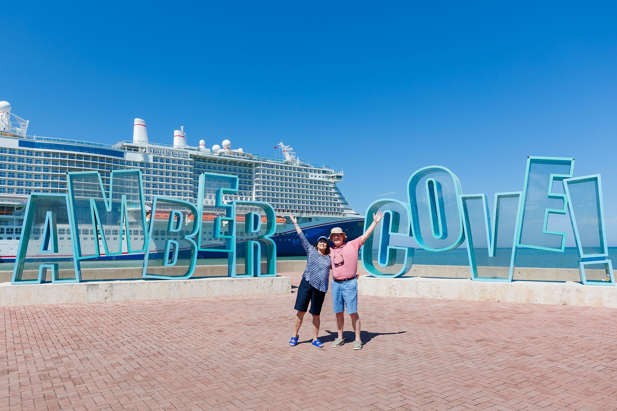 Two travelers stand with arms raised in front of the large turquoise "AMBER COVE" sign at the waterfront in the Dominican Republic. A cruise ship is docked behind them under a bright blue sky, creating a cheerful and welcoming port-of-call scene.