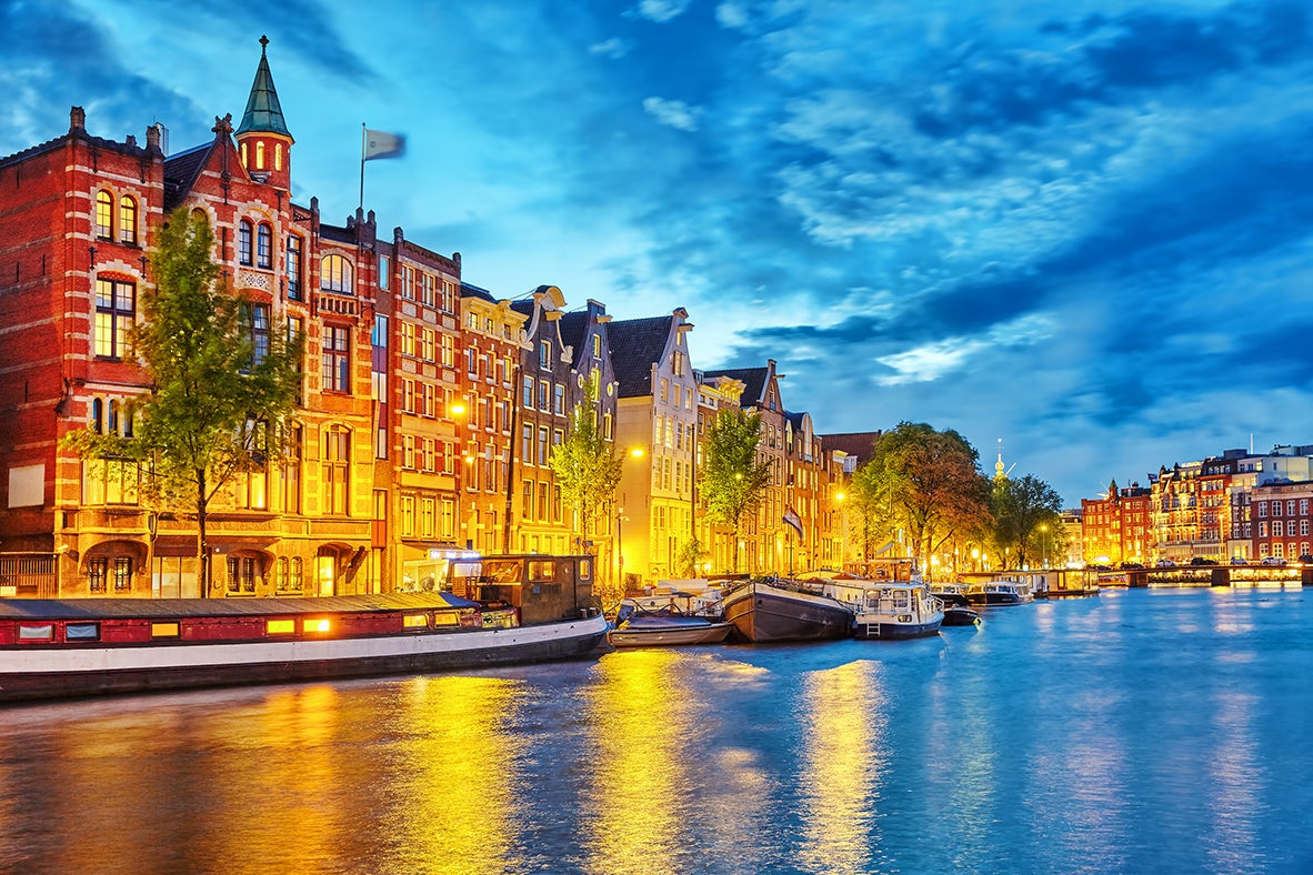 Amsterdam canal at dusk with historic canal houses, moored boats, and glowing city lights reflected on the water.