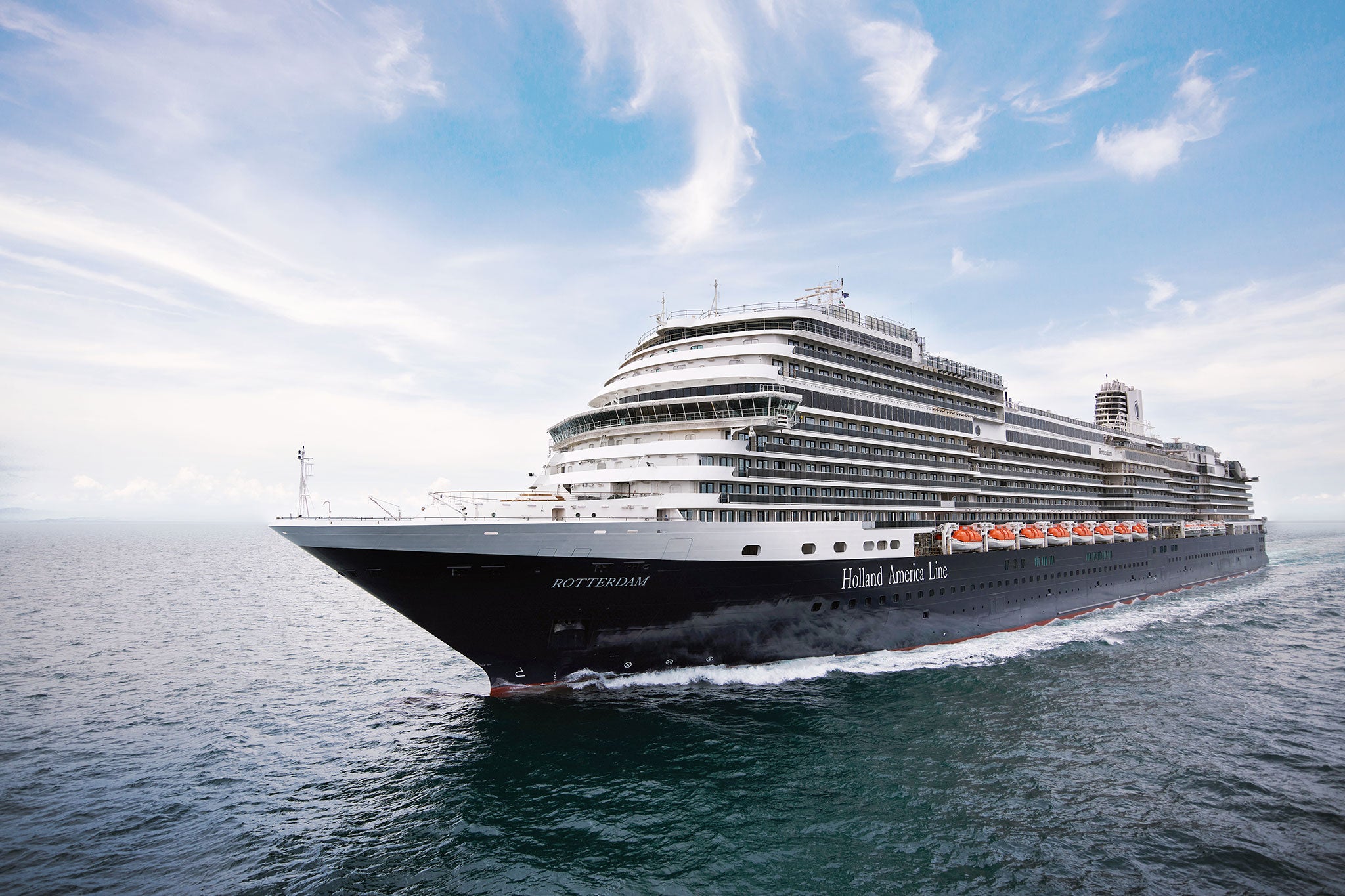 Holland America Line’s Rotterdam sails across open water beneath a bright blue sky streaked with wispy clouds.