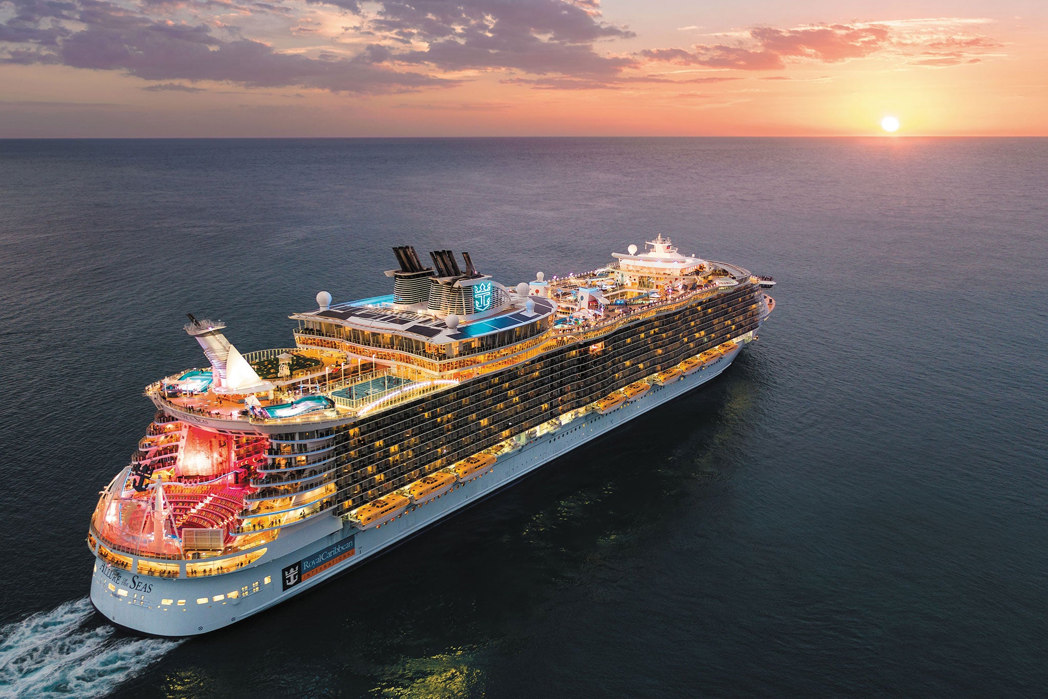 Royal Caribbean’s Allure of the Seas sails across calm ocean waters at sunset, its brightly lit decks glowing beneath pink and purple clouds.