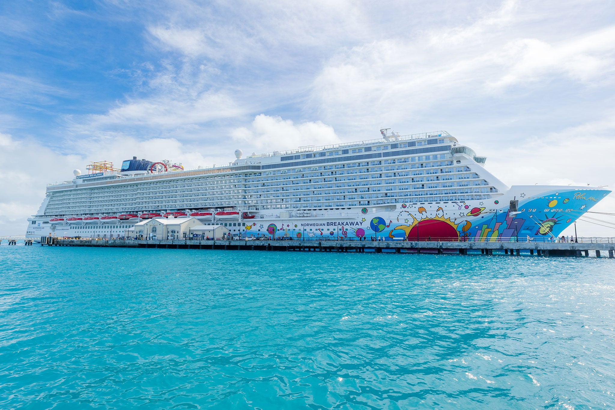 Norwegian’s Breakaway docked beside a pier, featuring colorful artwork along its hull and surrounded by bright turquoise water under a partly cloudy sky.