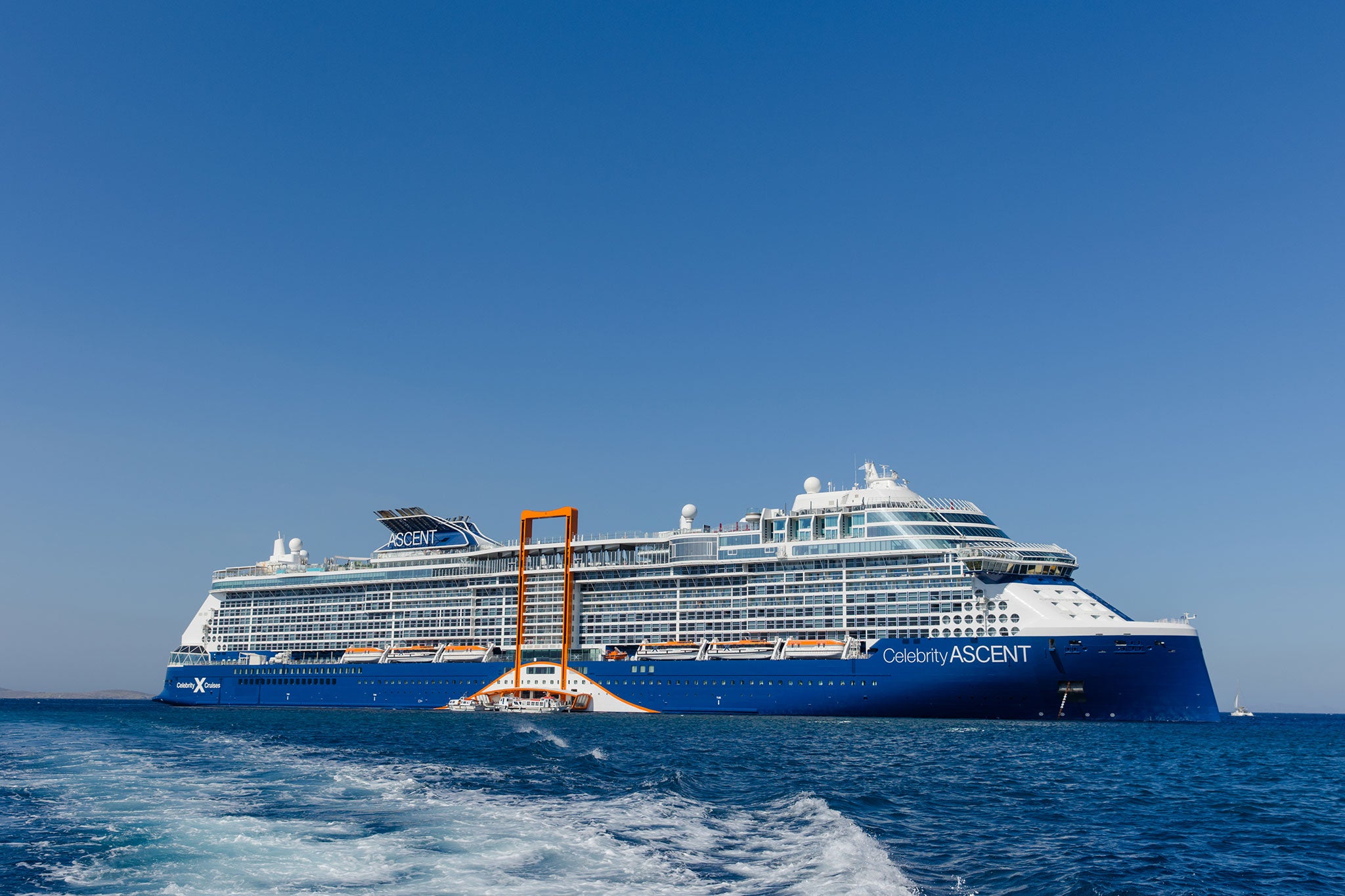 Celebrity Ascent sails across deep blue water beneath a clear sky, with the ship’s orange Magic Carpet platform extended along its side.