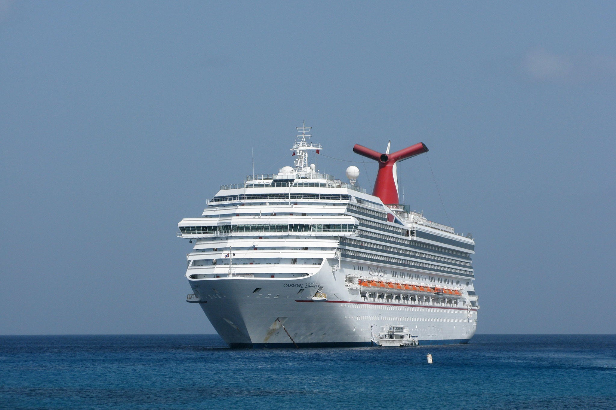 Large white cruise ship with a red funnel anchored on calm blue water beneath a clear sky.