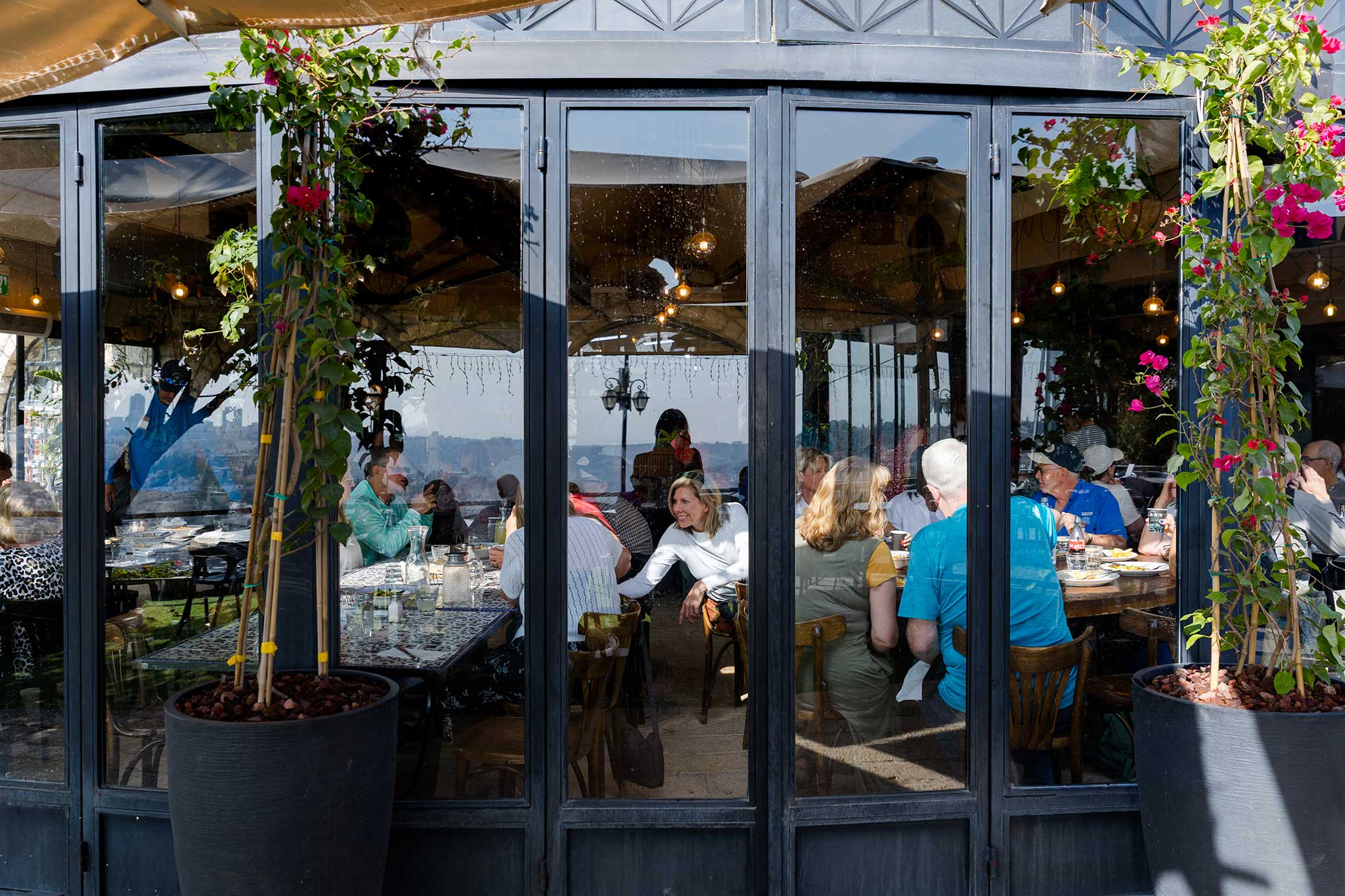 Travelers share a meal together inside a bright restaurant framed by large windows and colorful flowers.